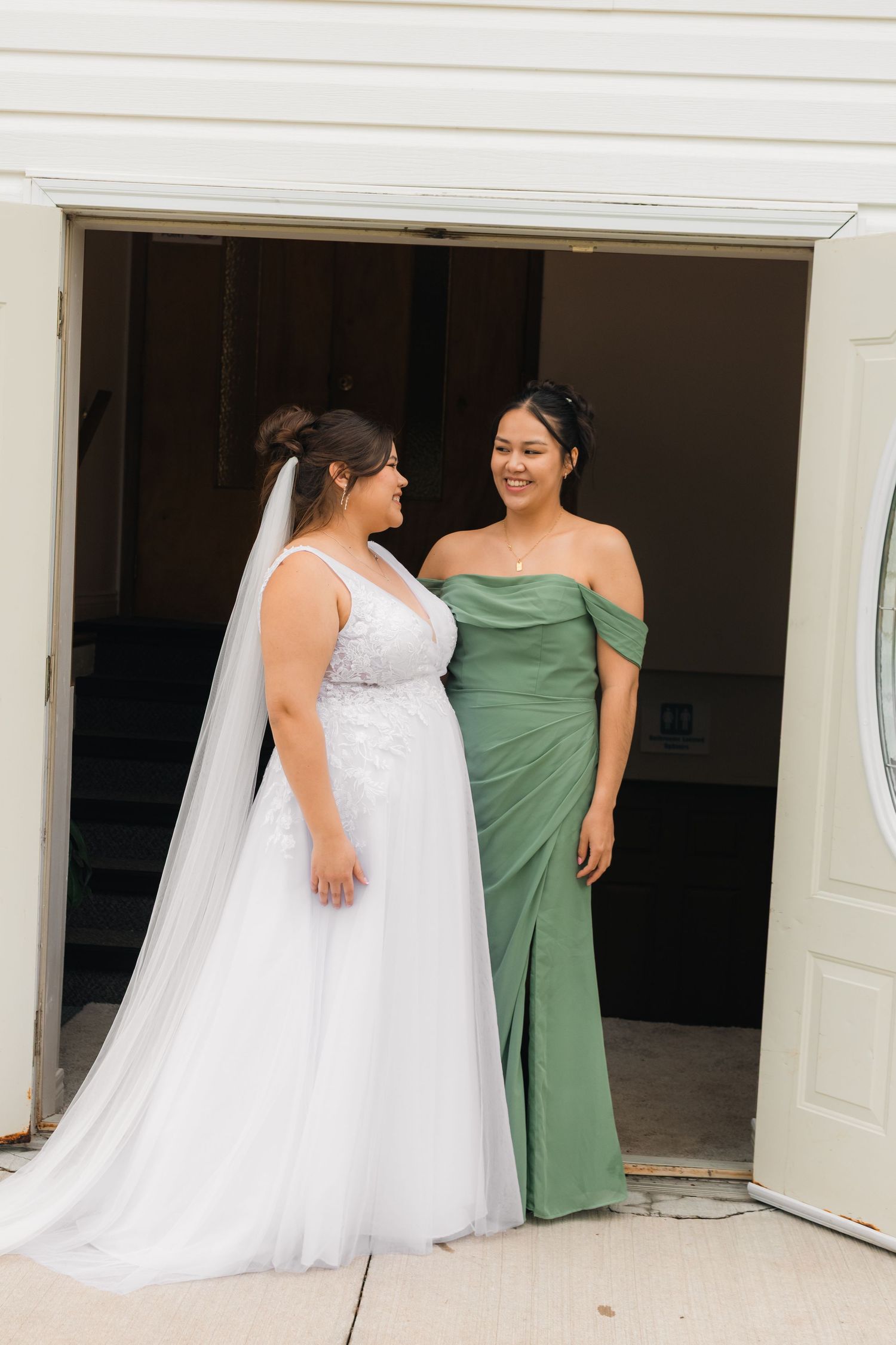 Bride in white lace gown and veil stands with bridesmaid in sage green off-shoulder dress in church doorway.