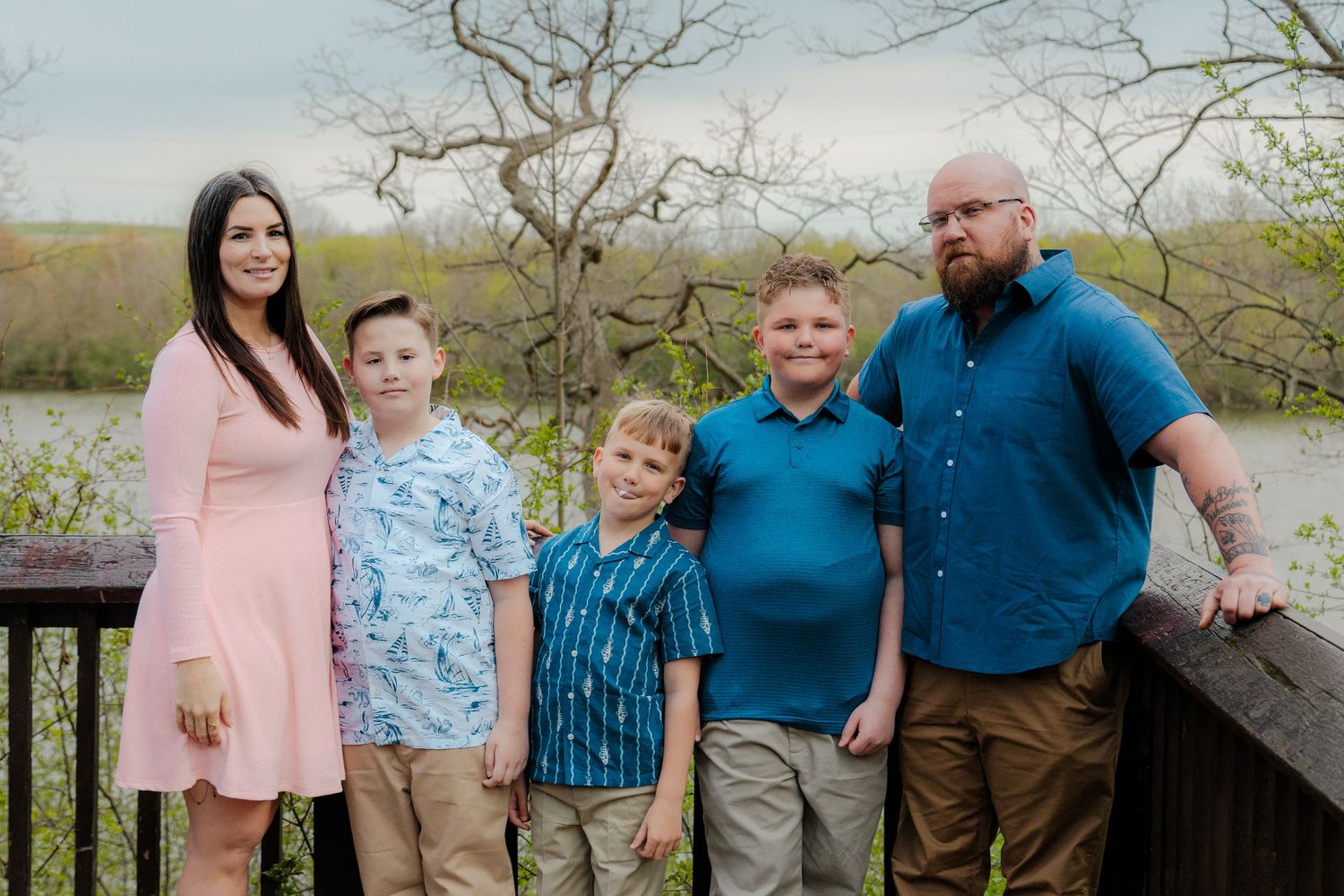 Family of five posing on wooden deck outdoors, with lush greenery and water in background on a spring day.