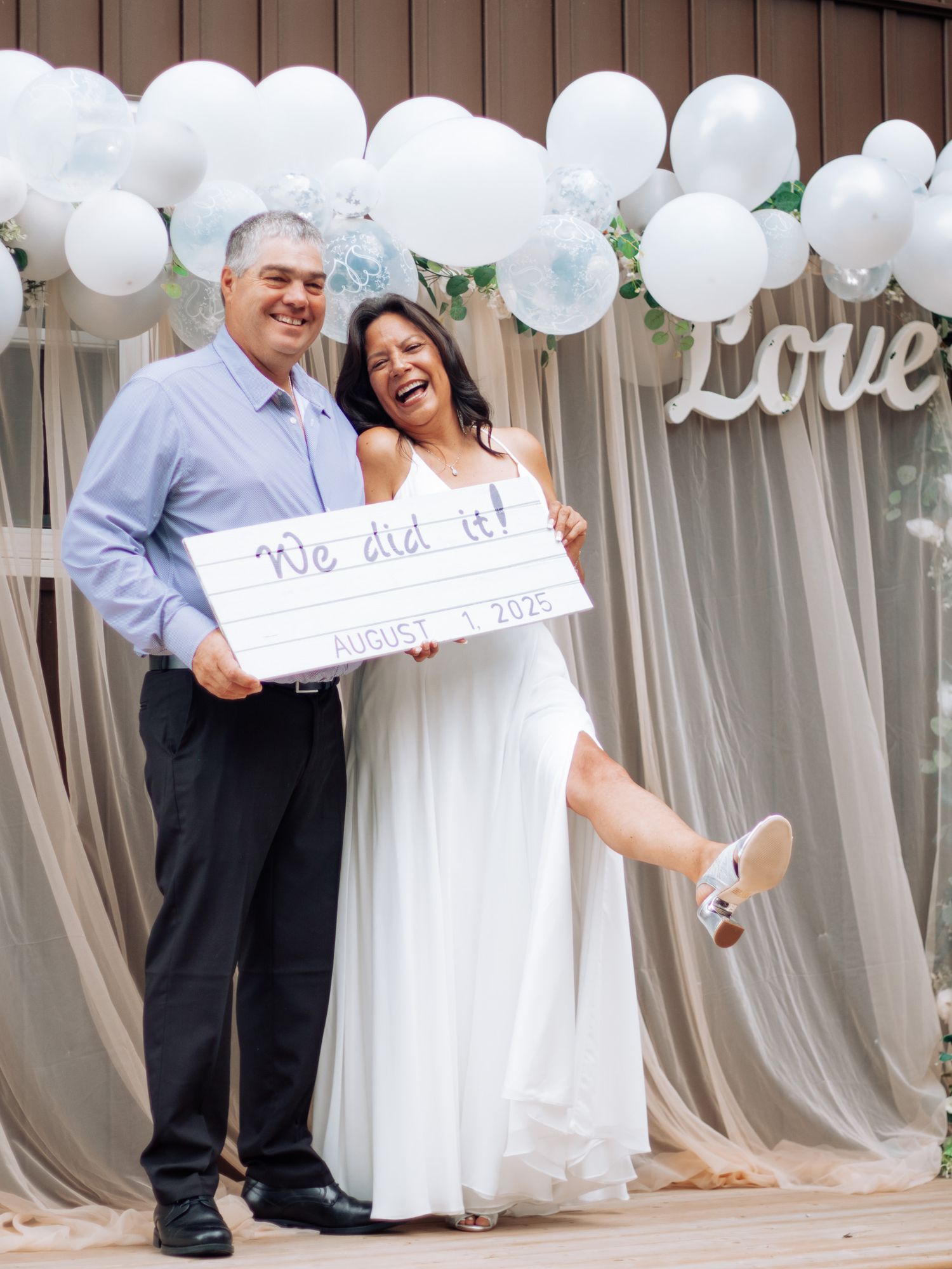 Newlyweds laughing and holding We Did It sign in front of balloon arch wedding backdrop.