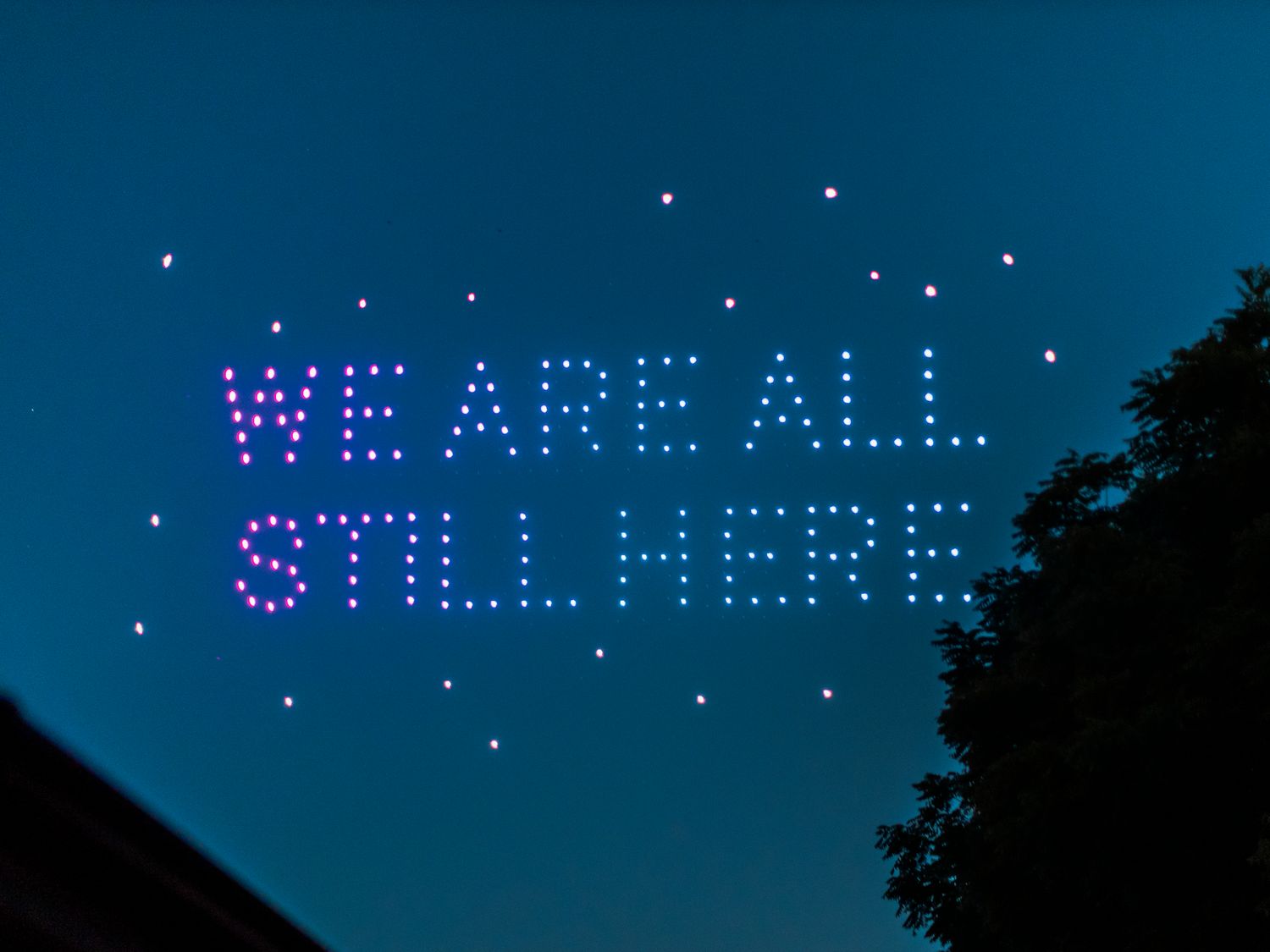 Drone light show spelling WE ARE ALL STILL HERE in pink LED lights against a twilight blue sky.