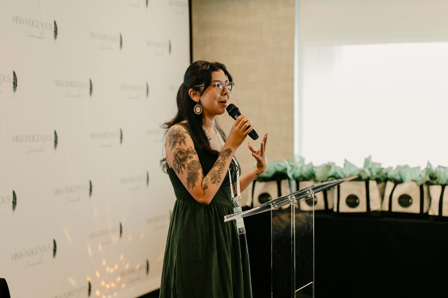 Woman with tattoos speaking at a podium during an indoor event, wearing a green dress and gold earrings.