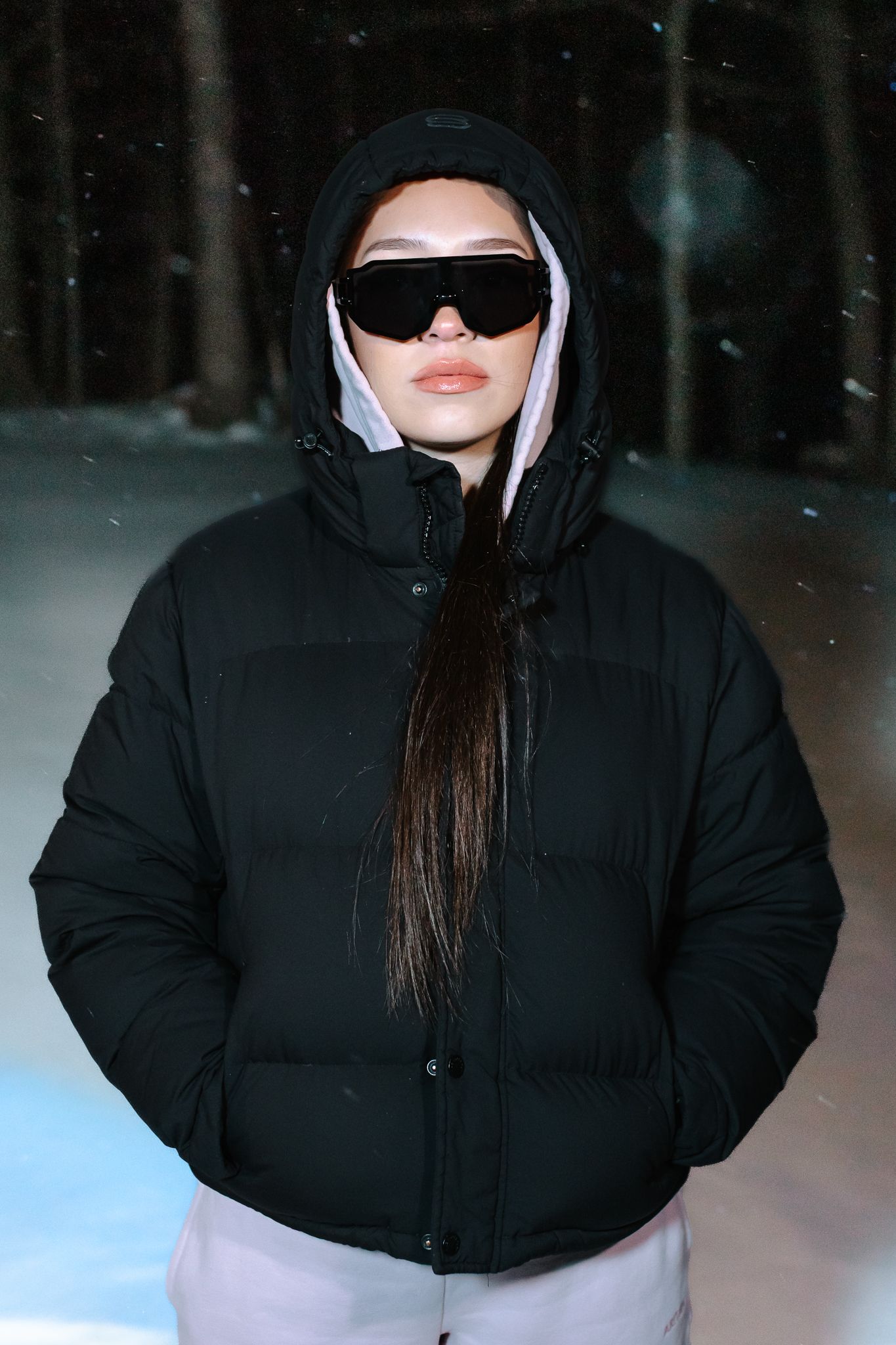 Woman in black hooded puffer jacket and sunglasses standing in a snowy forest at night.