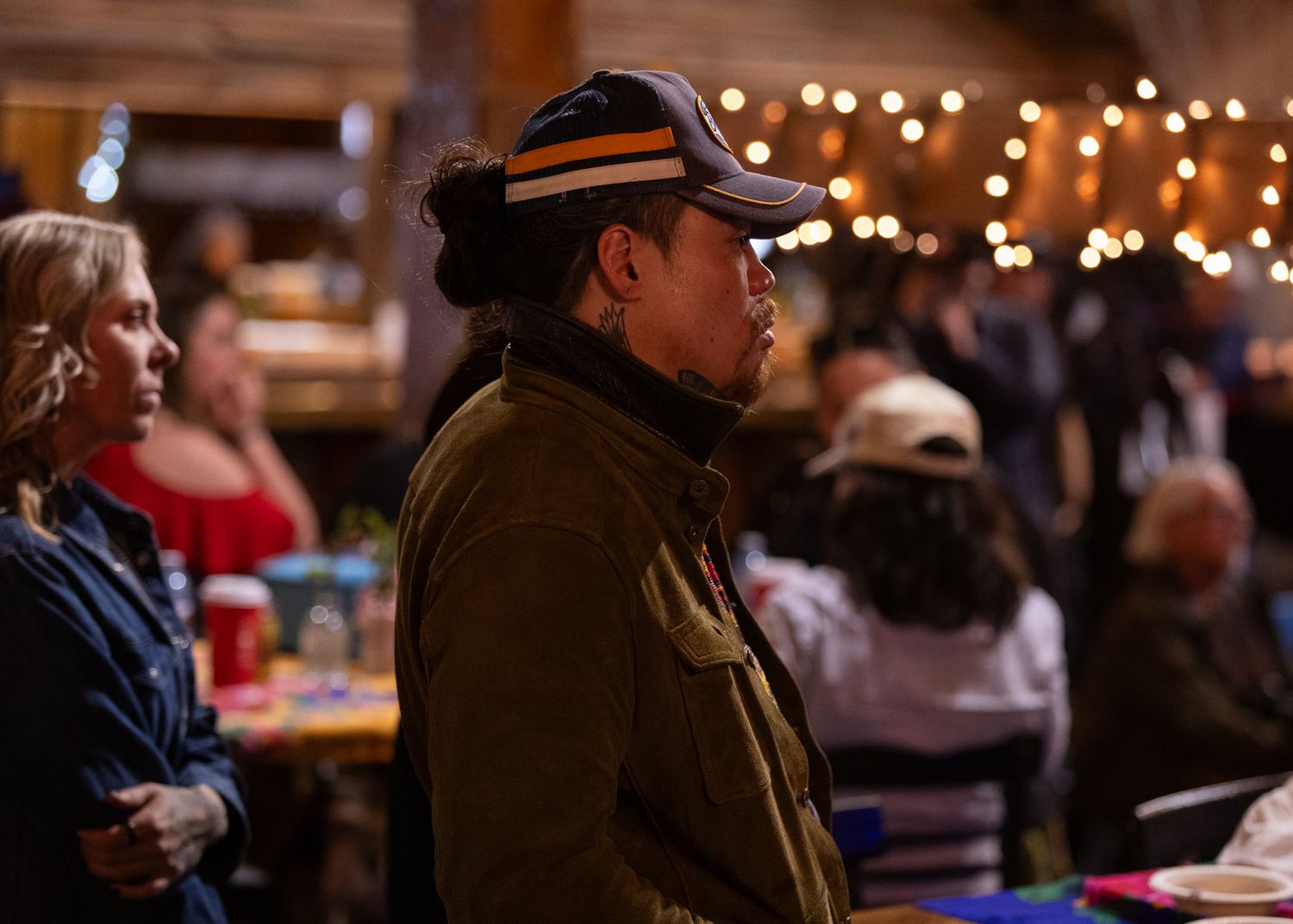 A person wearing a cap listens attentively at an indoor event with warm bokeh lights in the background.