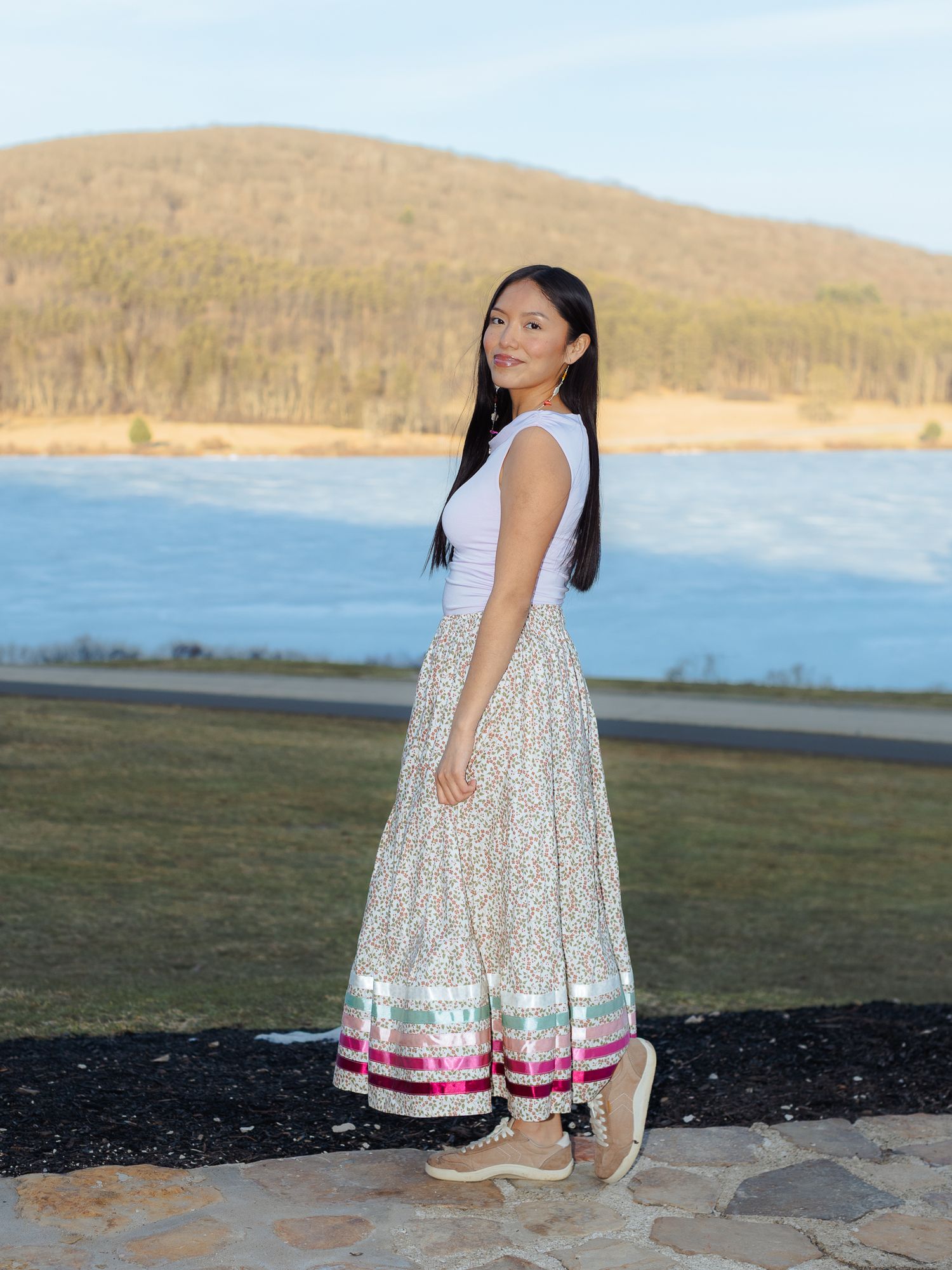 Young woman in white top and floral maxi skirt with pink trim poses by a scenic lake and mountain backdrop.