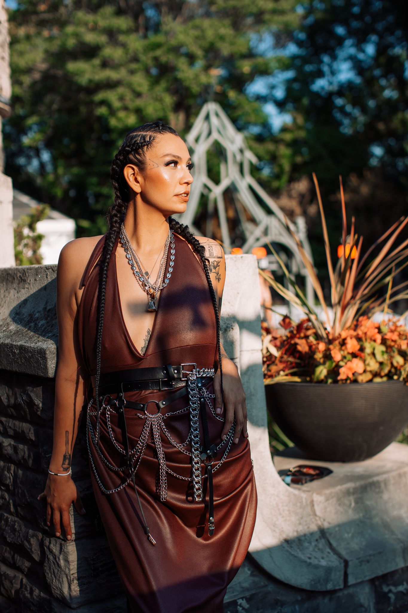 Woman in brown leather halter dress with chain belt and silver necklaces, posing outdoors with tattoos visible.