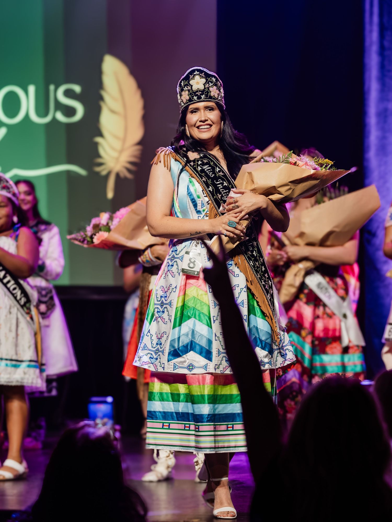 Indigenous pageant winner smiles on stage wearing traditional regalia, crown, and sash while holding a bouquet.