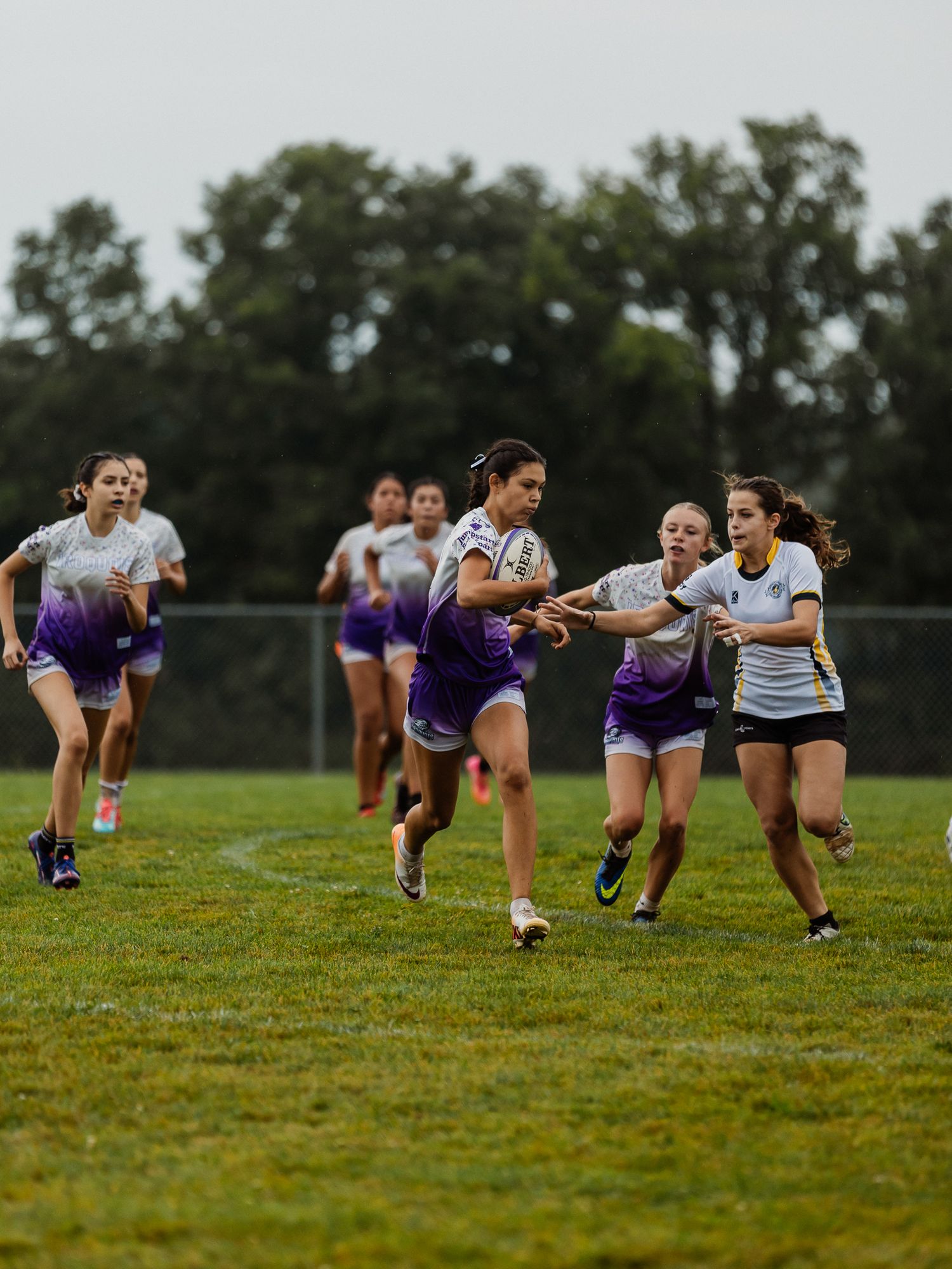 Female ultimate frisbee players in purple and white uniforms compete on a grass field during an outdoor tournament.