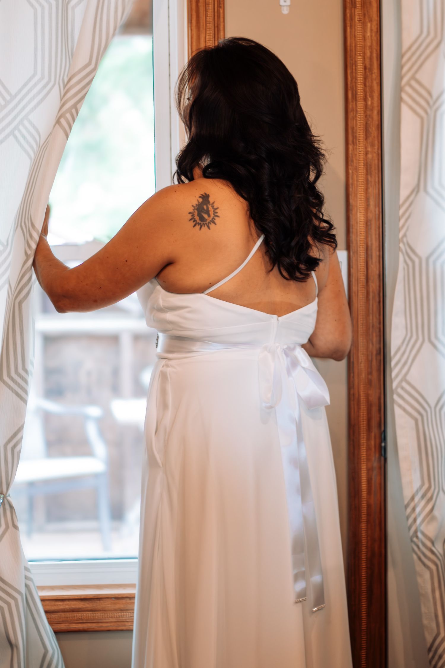 Bride in white wedding gown with satin bow, showing sun tattoo on upper back, standing by window with curtains.