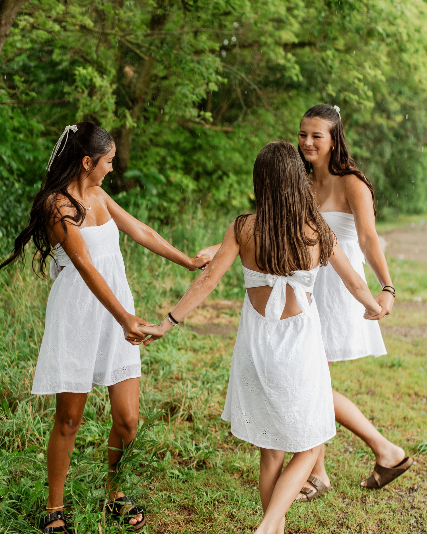 Three girls in white summer dresses hold hands and spin together in a green outdoor meadow setting.