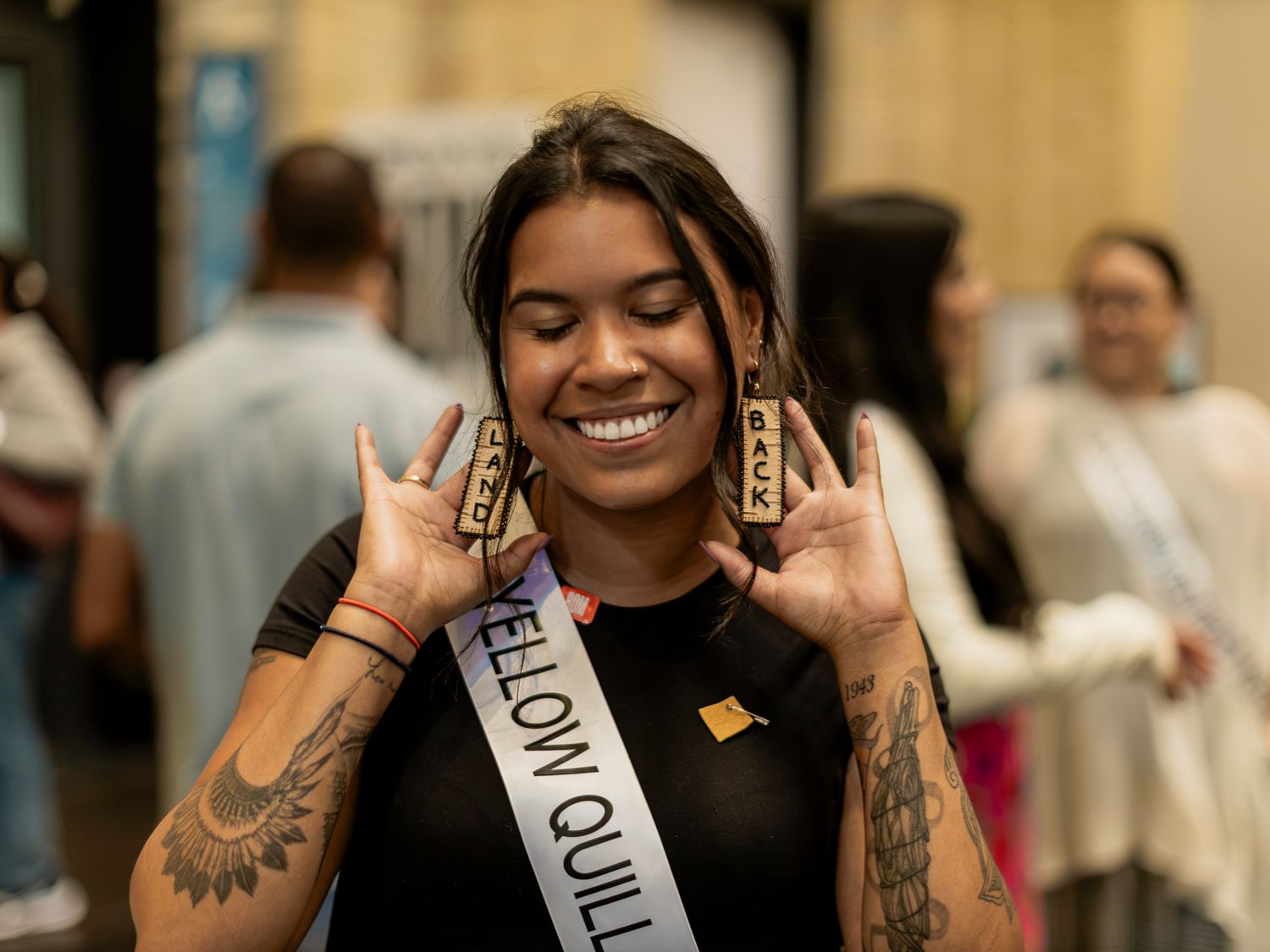 Young Indigenous woman wearing a Yellow Quill sash smiles while showing earrings.