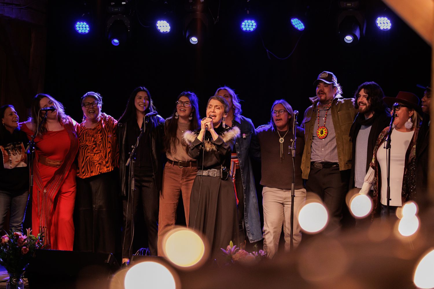A large group of performers stand together on a colorful stage during a musical ensemble finale.