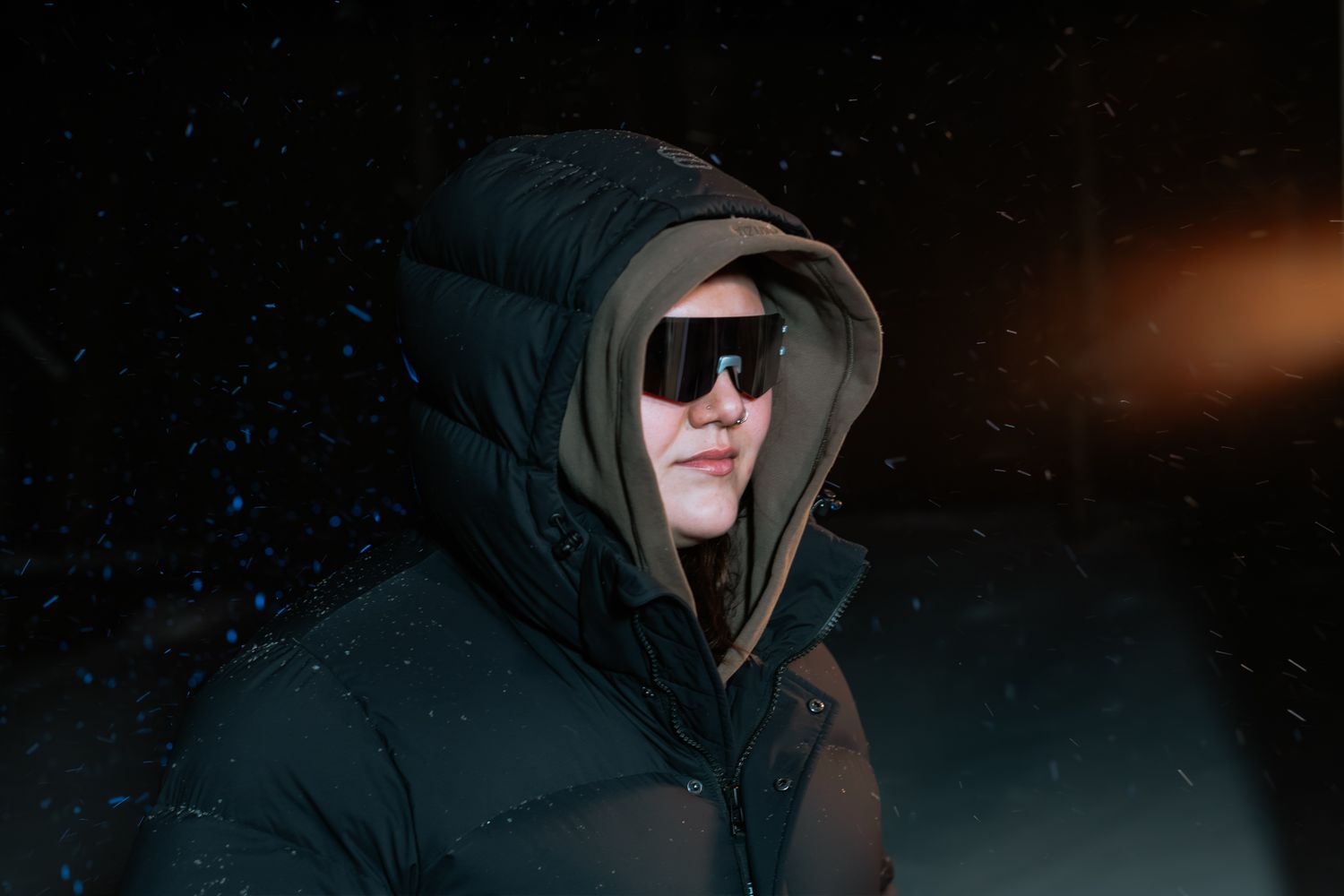 Person wearing black hooded puffer jacket and shield sunglasses against a dark snowy night sky.