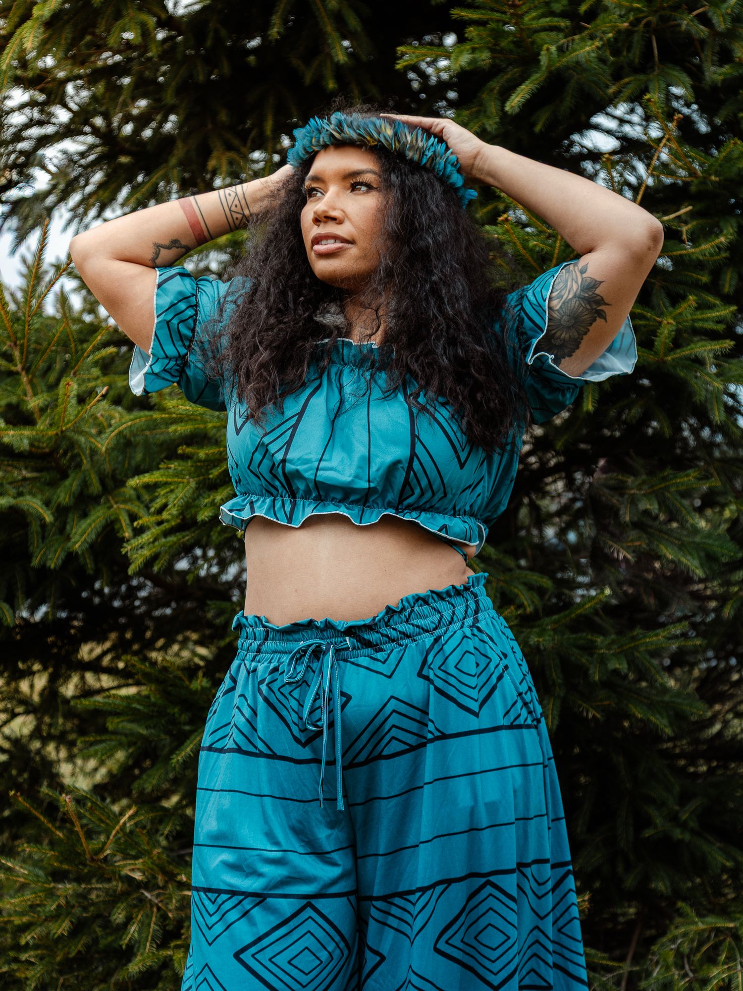 Woman in teal geometric print matching crop top and wide-leg pants set, posing outdoors near evergreen trees.