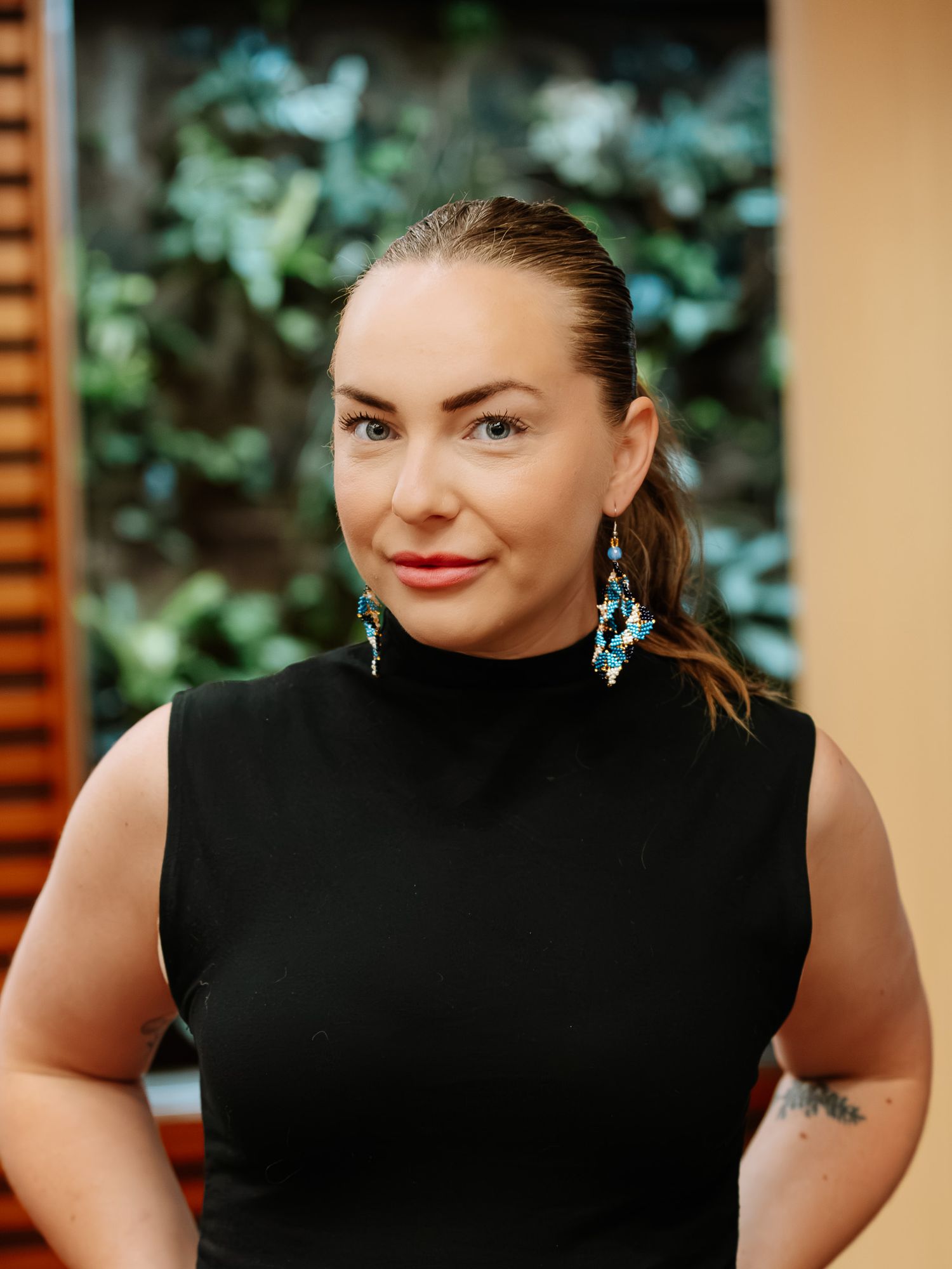 Woman in black sleeveless turtleneck with turquoise earrings and tattoo, posing against green foliage backdrop.