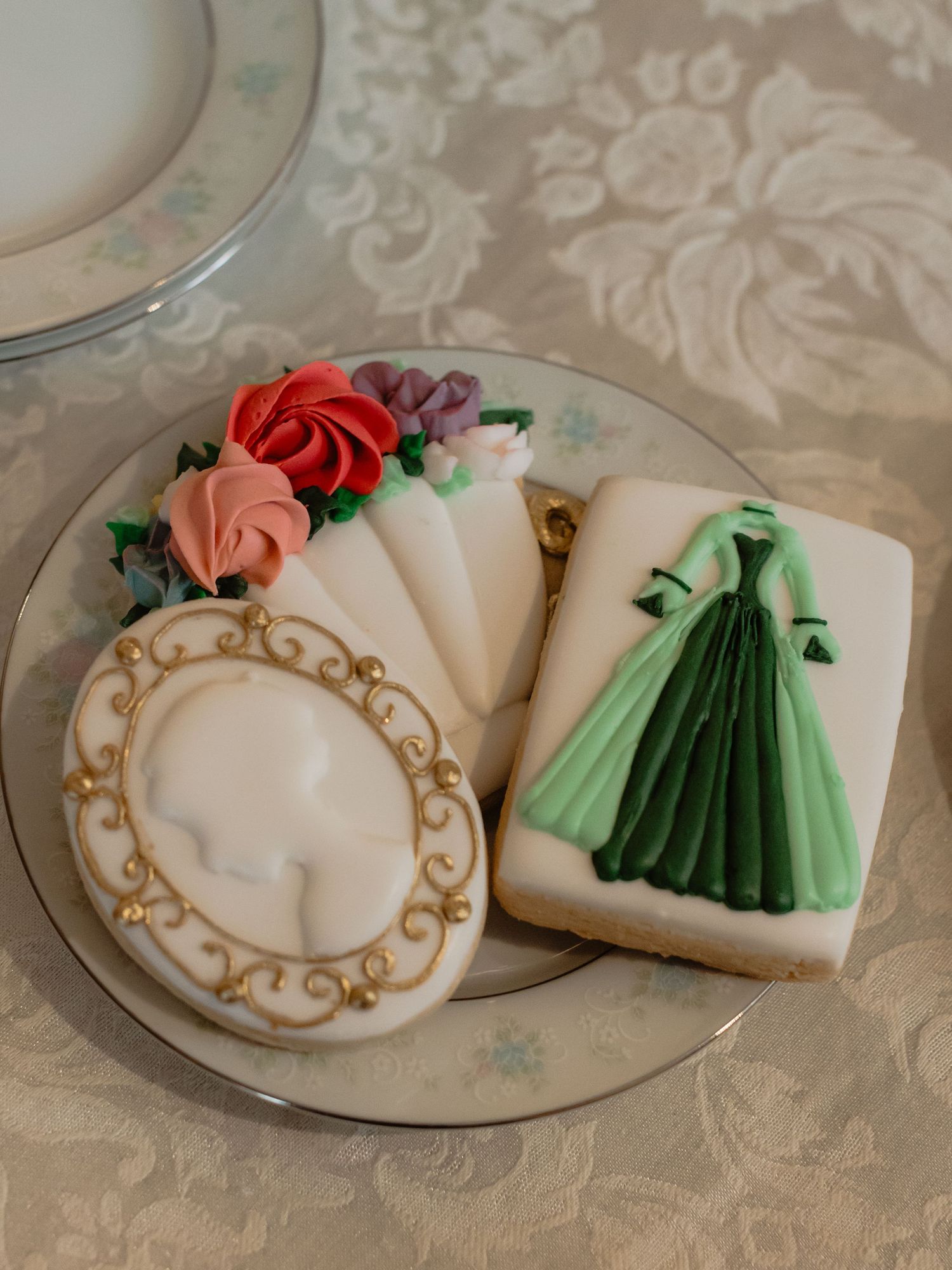 Elegant decorated sugar cookies featuring a green ball gown, floral bouquet, and ornate cameo frame design on a vintage plate.
