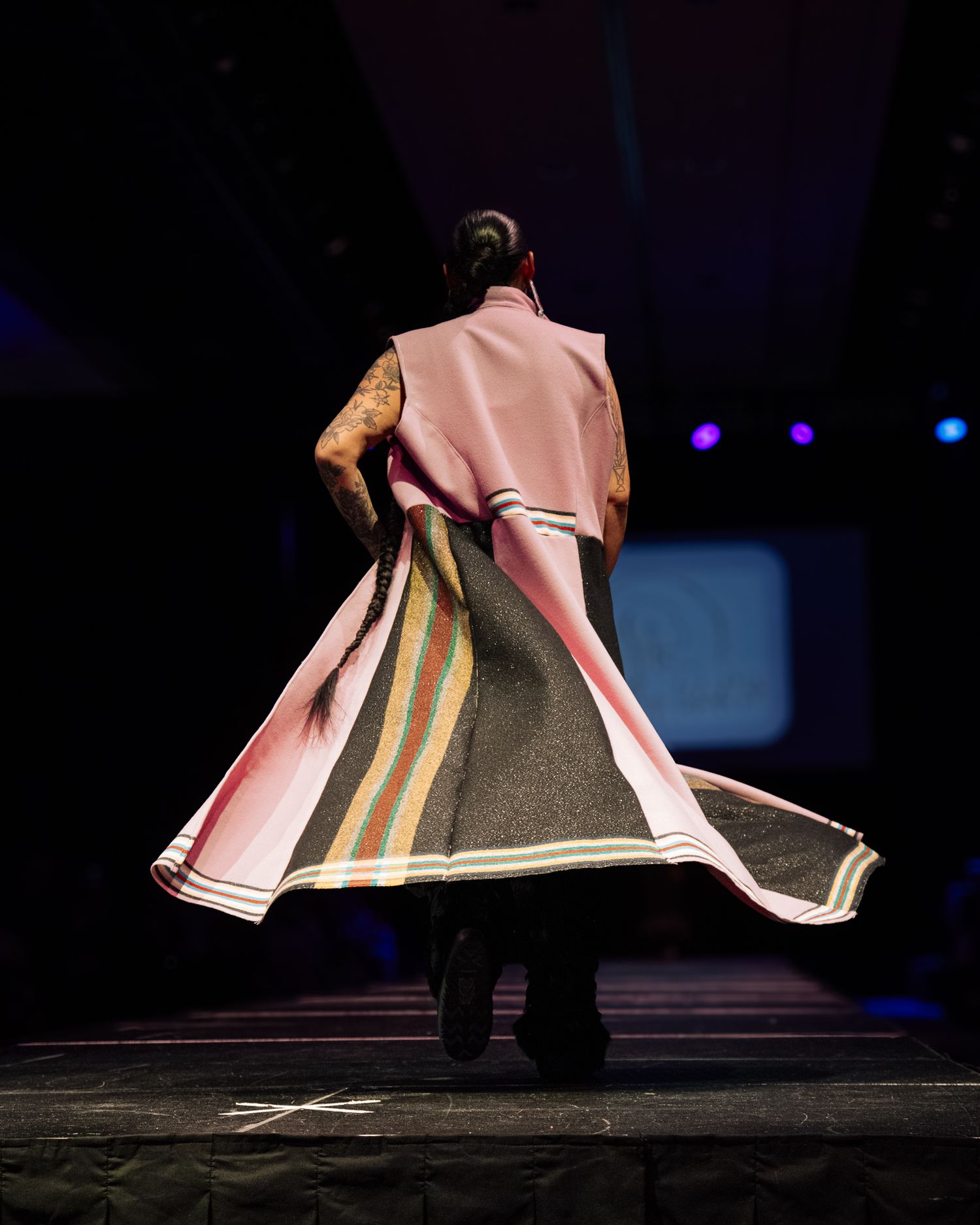 Model walks runway in flowing pink and gray striped coat, viewed from behind under dramatic stage lighting.