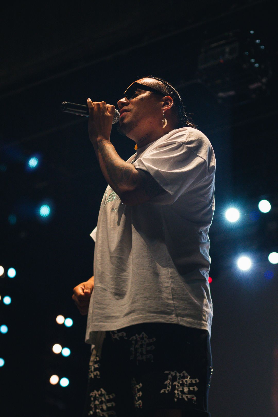 Rapper performing on stage with microphone, wearing white tee and sunglasses, with colorful stage lights behind.