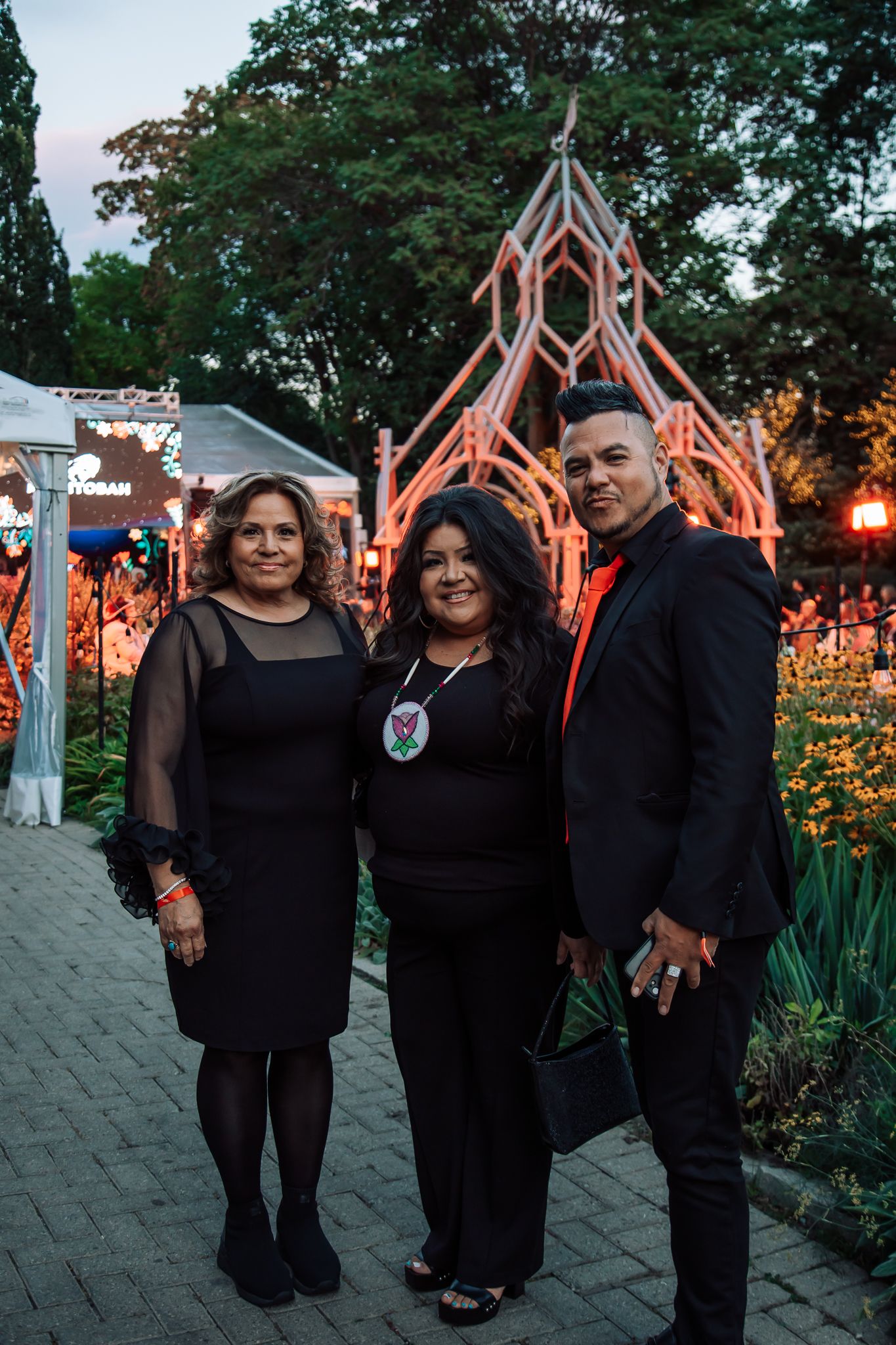 Three people in black formal wear pose outdoors at a nighttime event near a glowing geometric art installation.