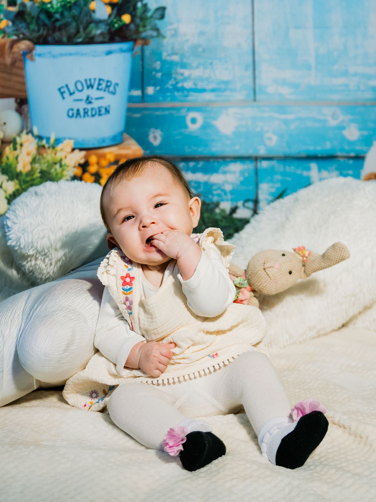 Adorable baby in white outfit sits on fluffy blanket near Flowers and Garden sign with stuffed bunny.