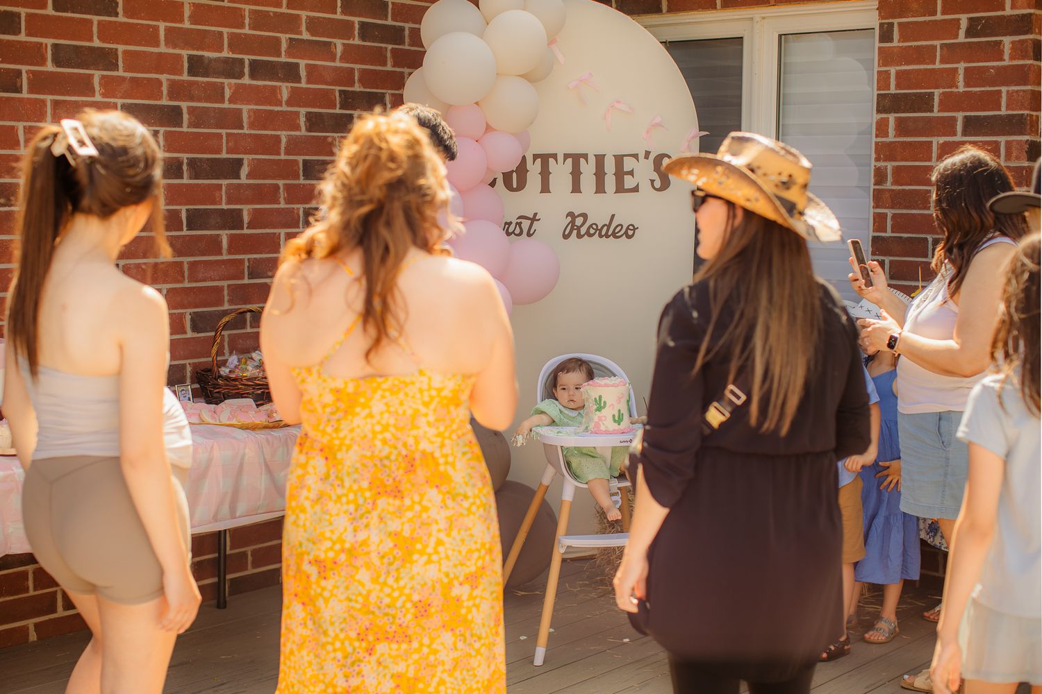 Guests gather around a high chair at a baby's first rodeo birthday party outdoors.