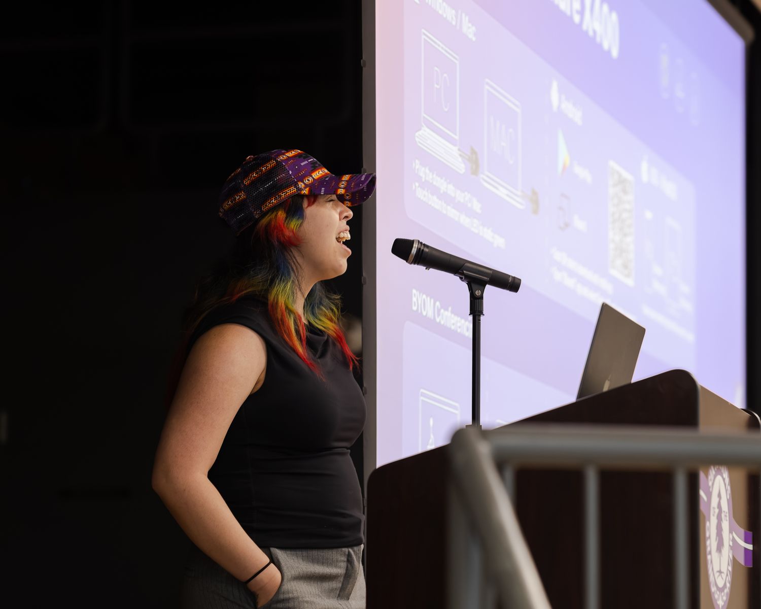 Young woman with colorful hair presenting at a podium with a large screen behind her.