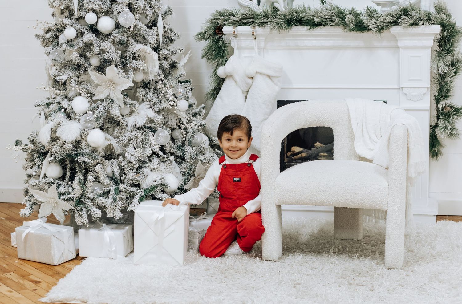 A child in red pajamas plays near a decorated Christmas fireplace and flocked tree.