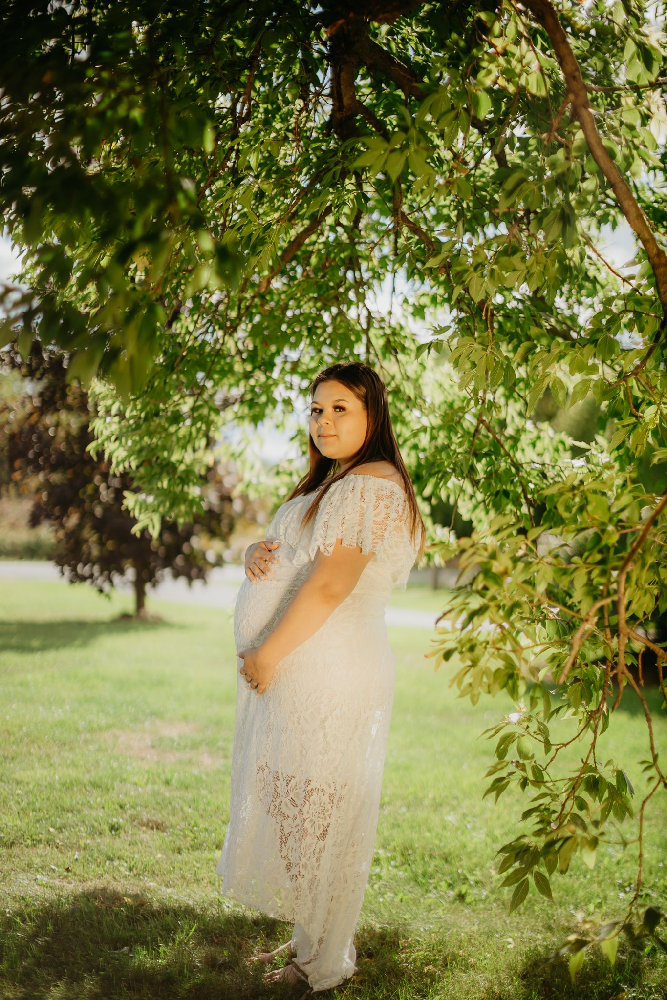 A maternity portrait of someone in a white lace dress standing under green tree branches.
