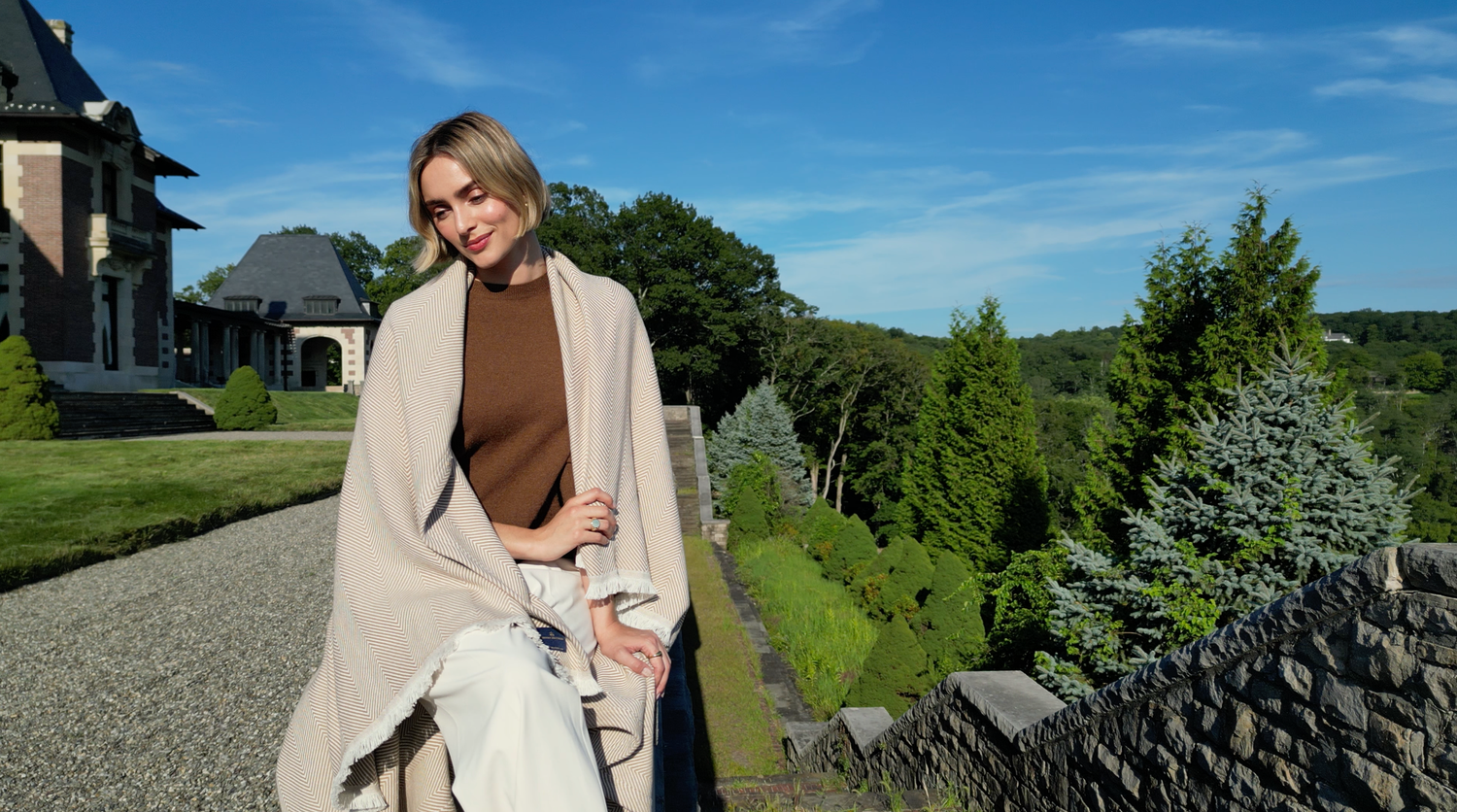 Woman in beige wrap and brown sweater smiling outdoors near stone path with lush green trees and elegant estate.