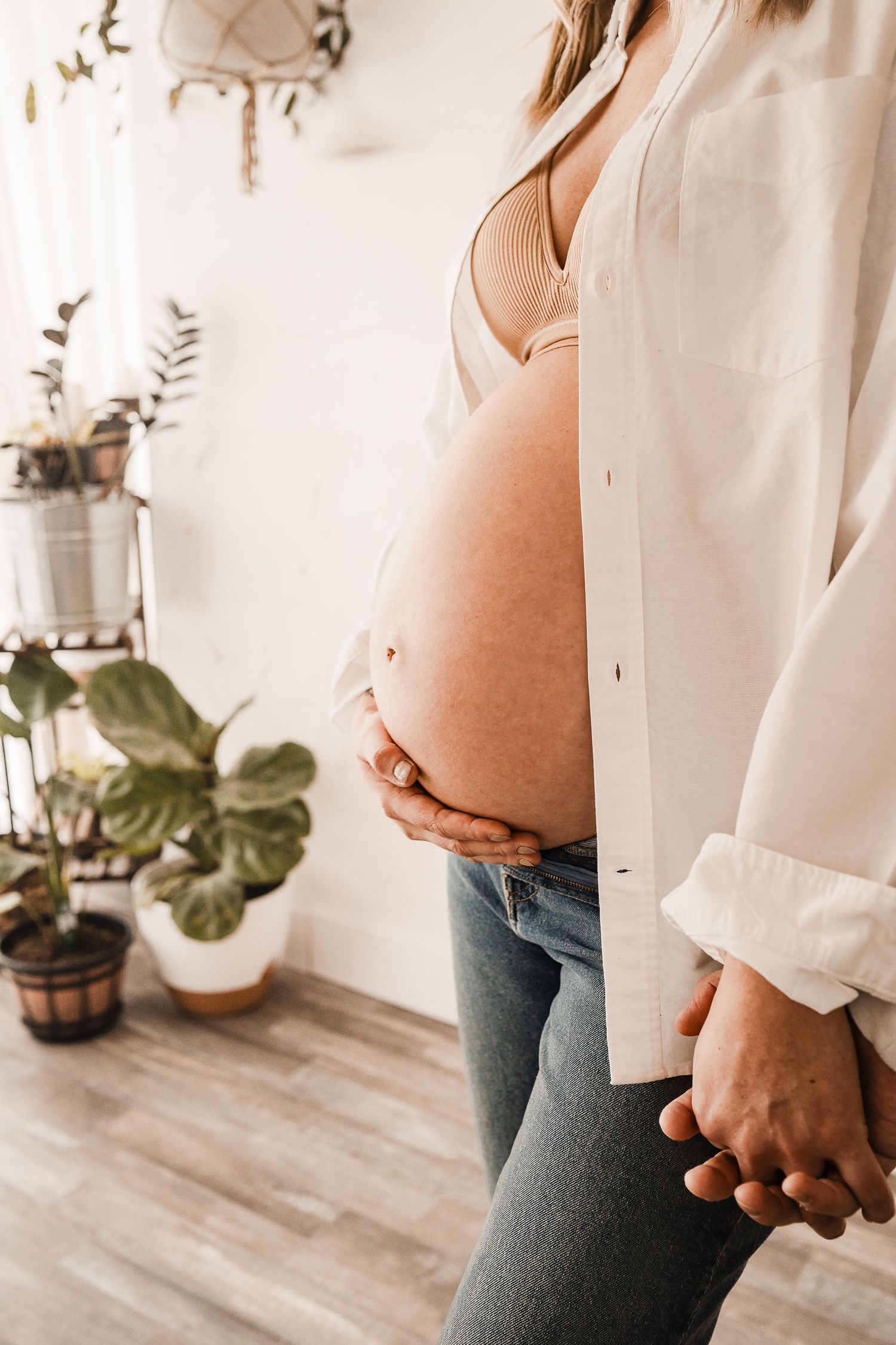 Pregnant woman in white shirt and jeans cradling her bare baby bump indoors near houseplants.