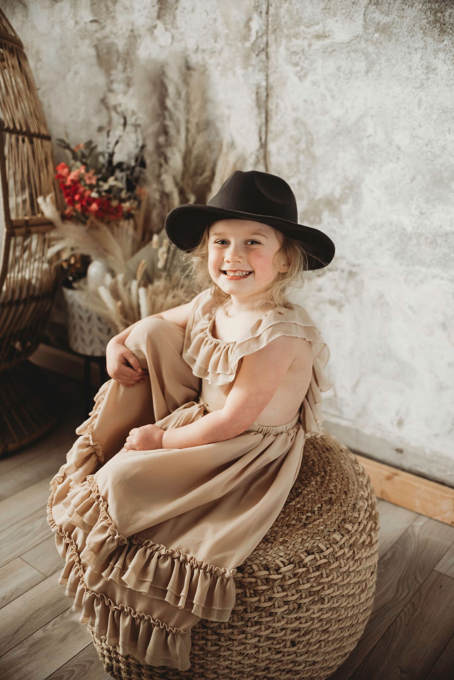 Young girl in beige ruffled dress and black hat sitting in a wicker basket chair.