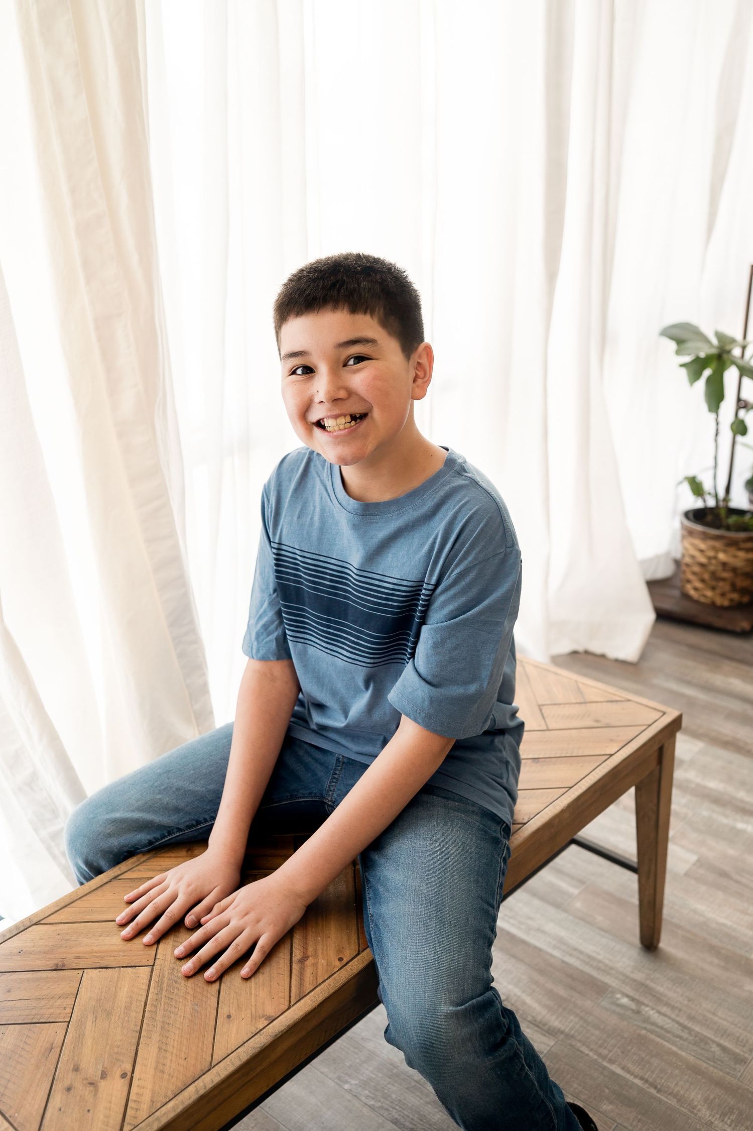 Smiling preteen boy sitting on a wooden bench in front of bright white curtains.