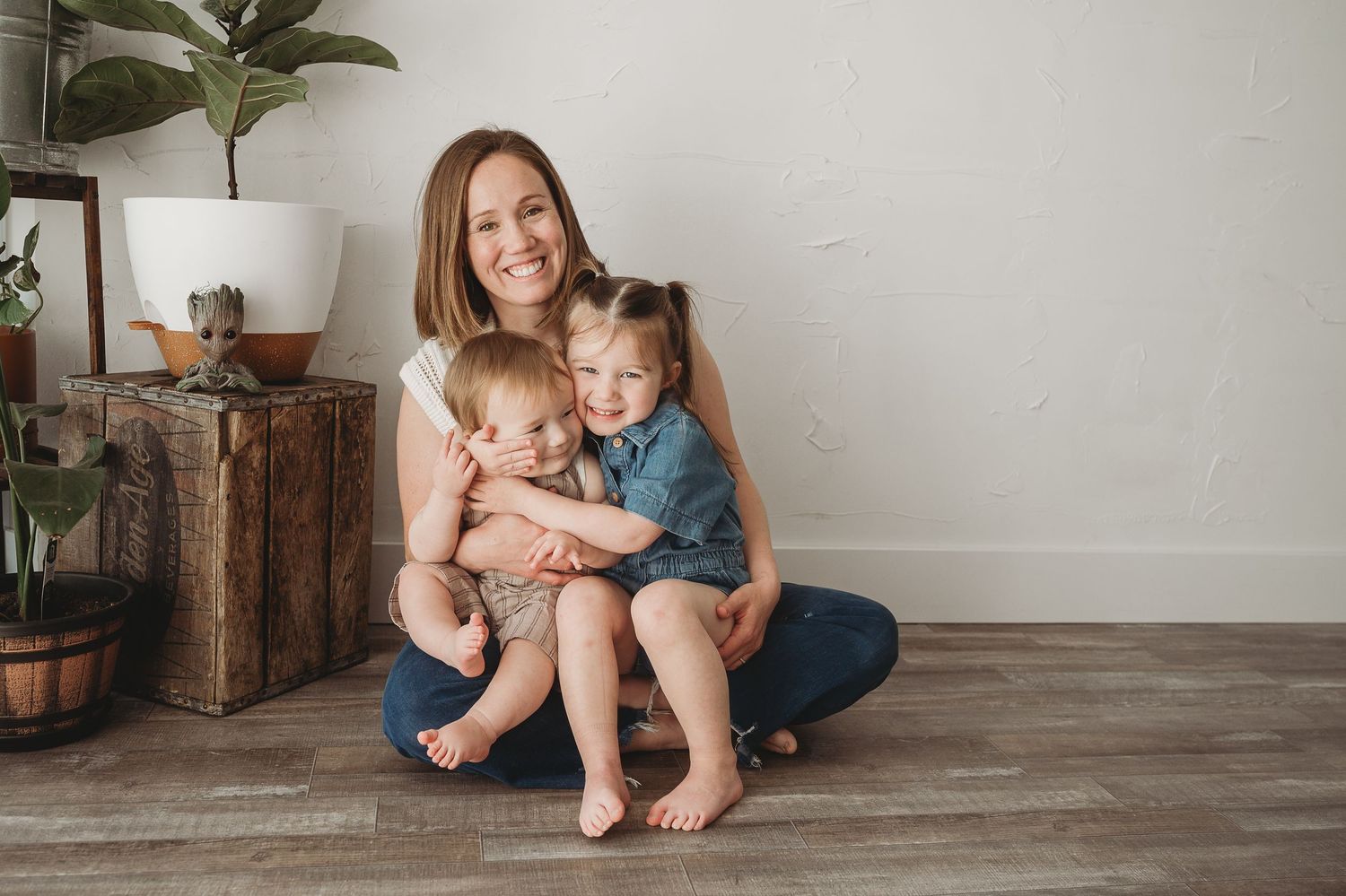 Smiling mother sitting on floor hugging two young children in a bright, plant-filled room.
