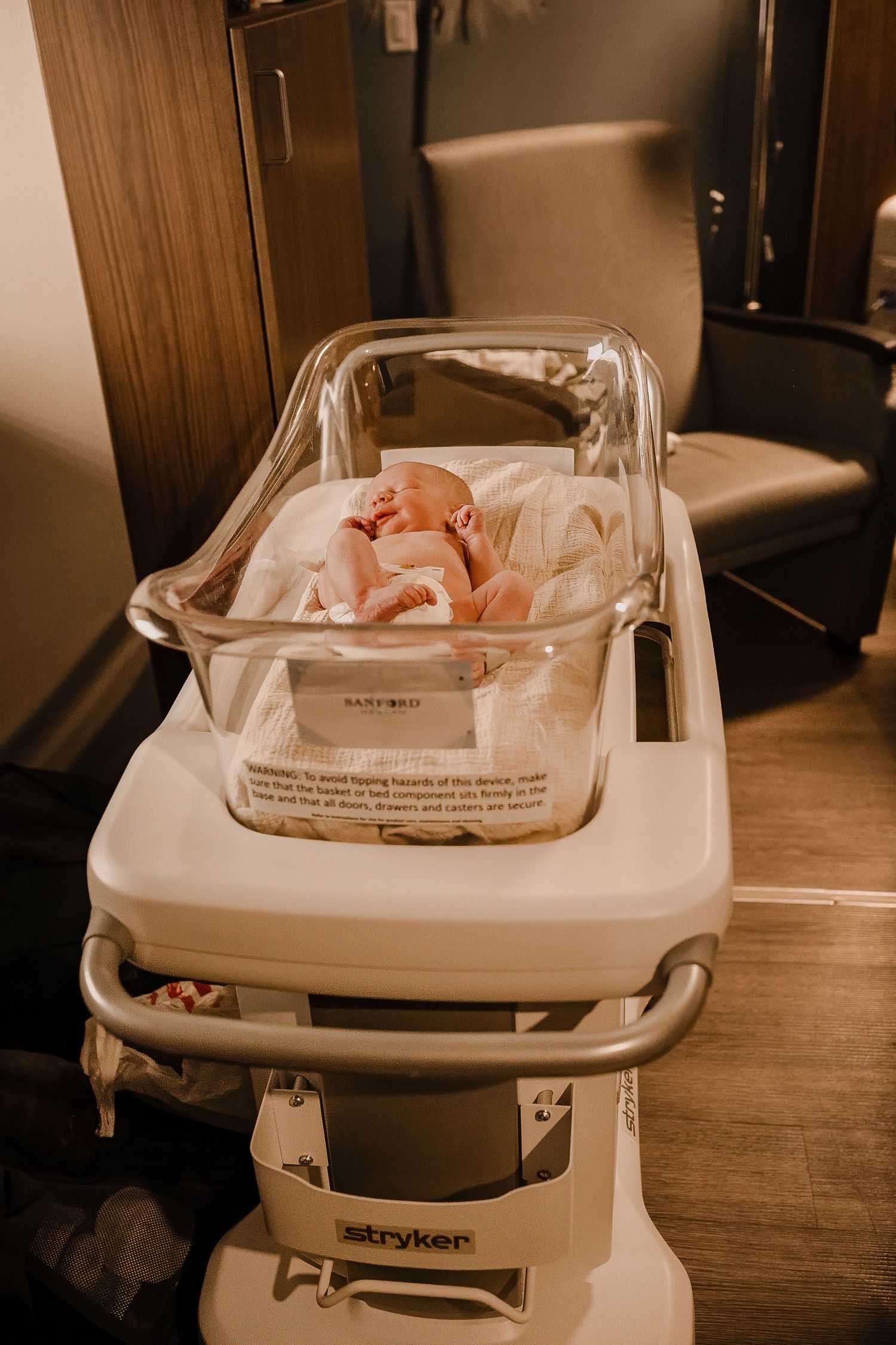 Newborn baby sleeping peacefully in a hospital bassinet on a Stryker medical cart.