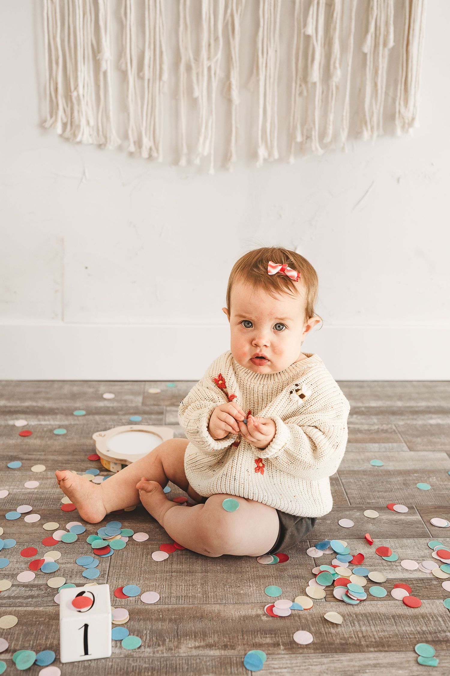 Baby sitting on wooden floor with colorful confetti during milestone photo session.