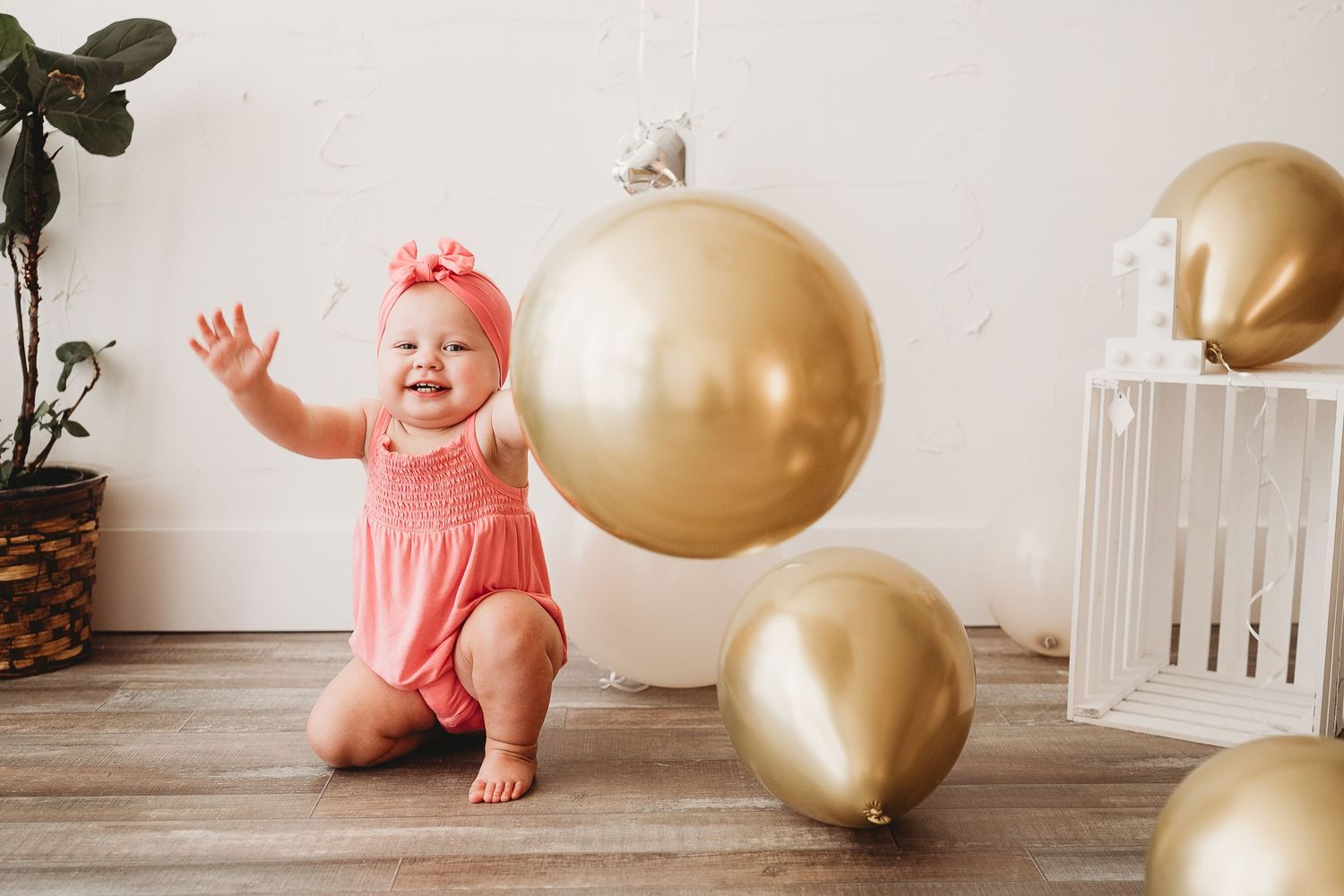 Baby in pink outfit sits on wooden floor surrounded by gold balloons and white crib for first birthday photoshoot.