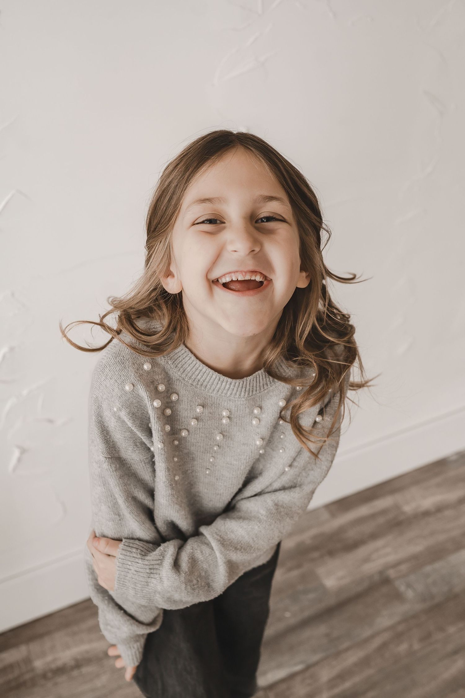 Joyful young girl in a grey pearl sweater laughing and leaning forward against a white wall.