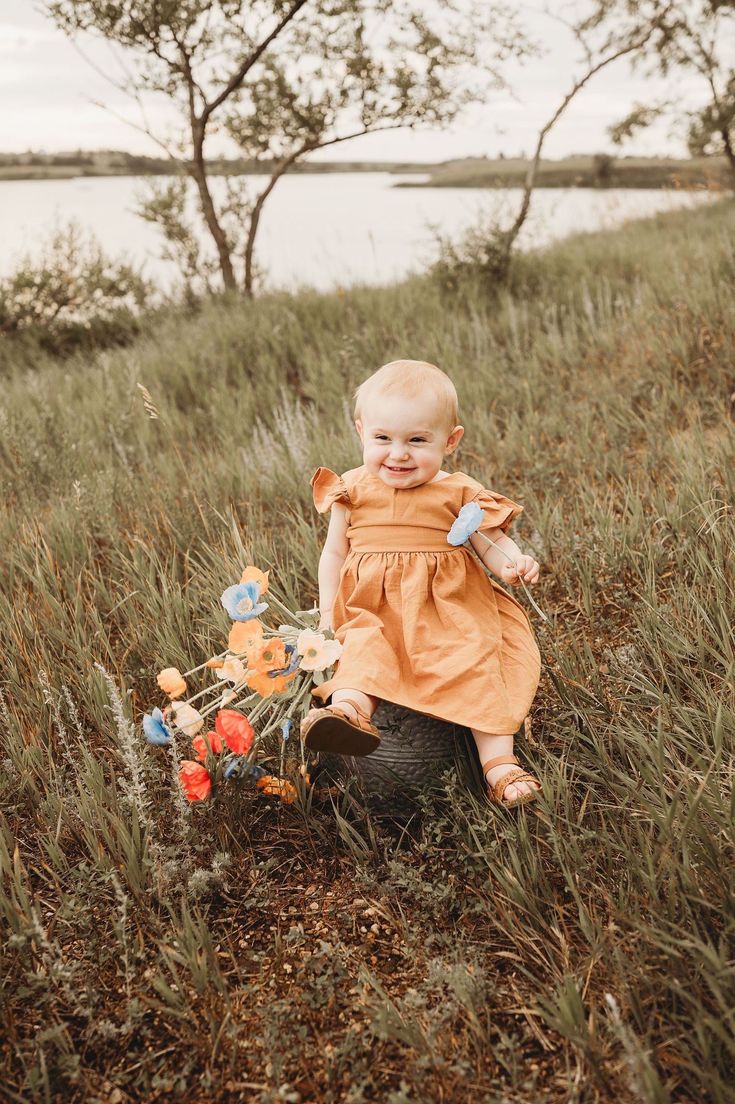Happy baby in orange dress sitting in a field with colorful flowers near a lake.