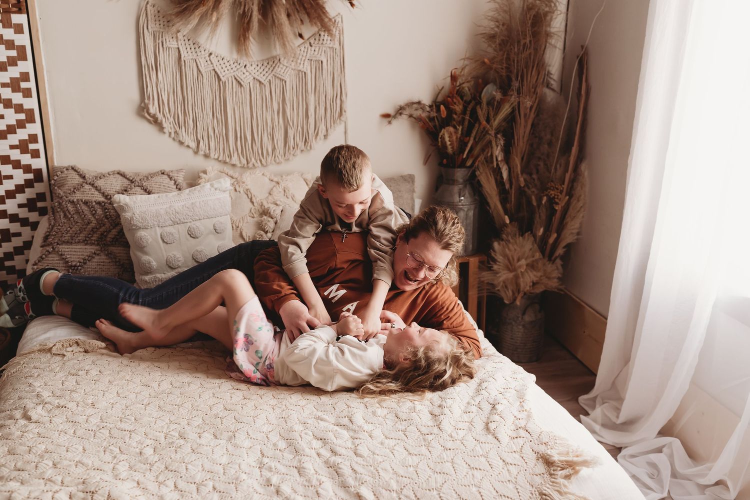 Three children playing and laughing together on a cozy bed in a bohemian decorated bedroom.