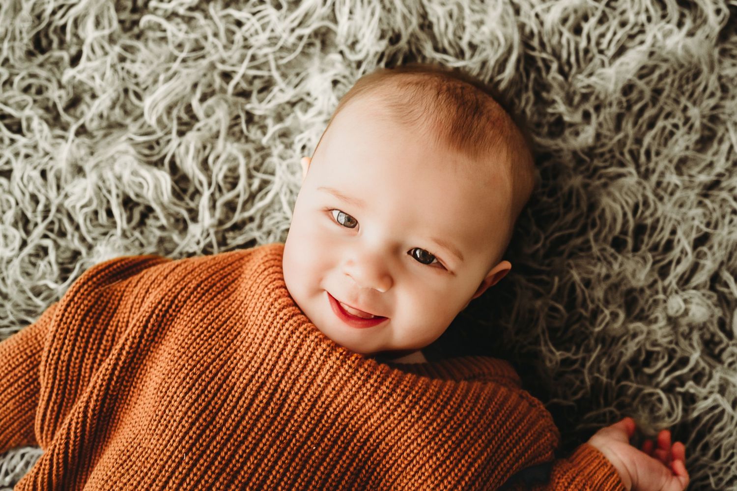 Smiling baby wearing burnt orange sweater lying on shag carpet.