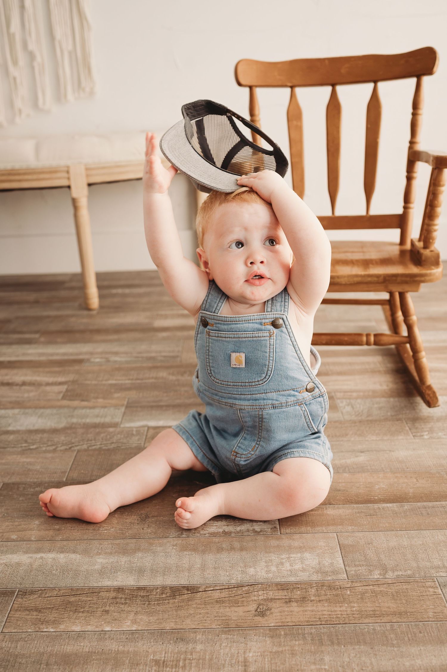 Baby in denim overalls playing with hat on wooden floor.