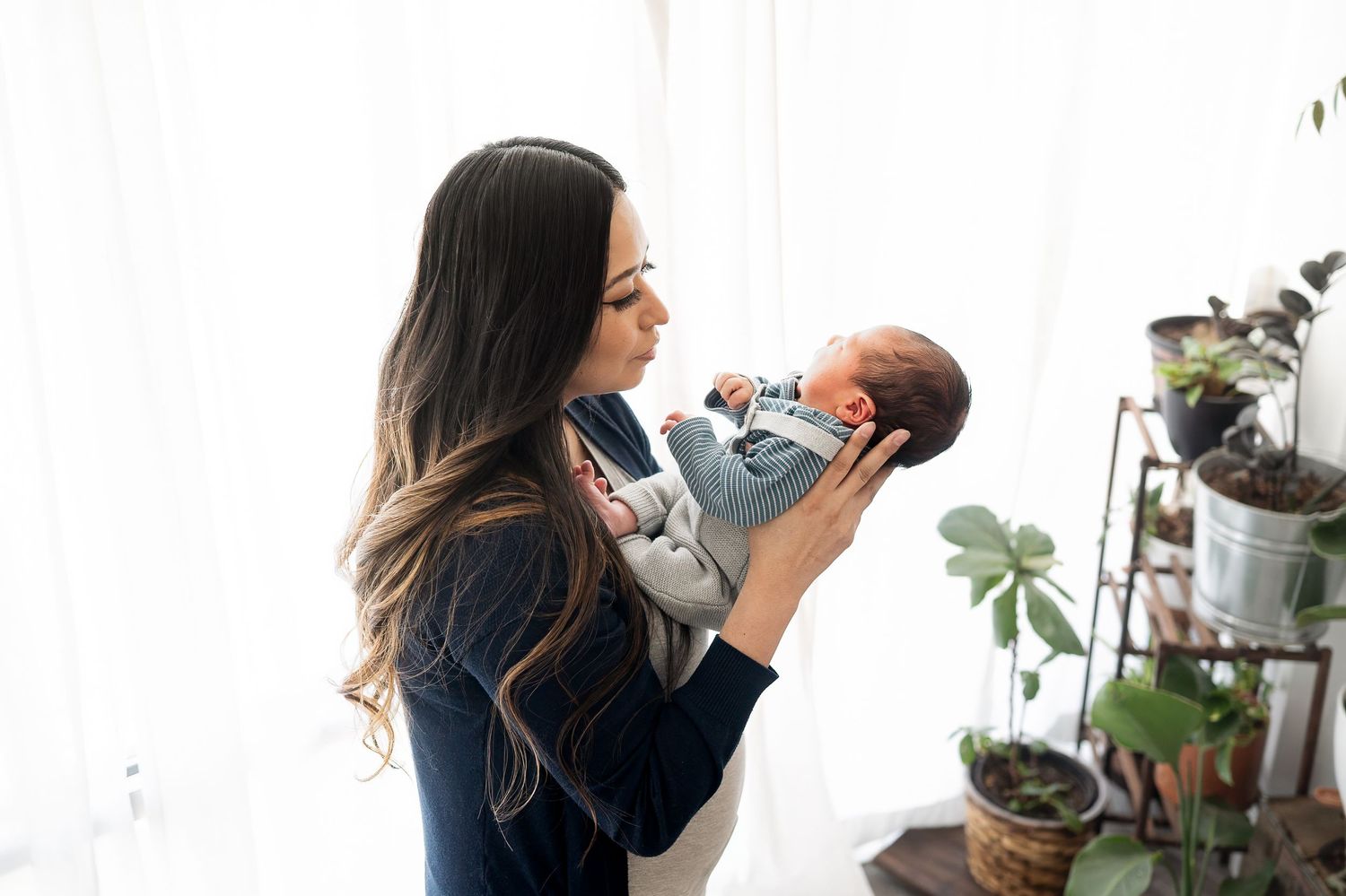 Dark-haired woman lovingly gazing at her newborn baby near a plant shelf by a window.