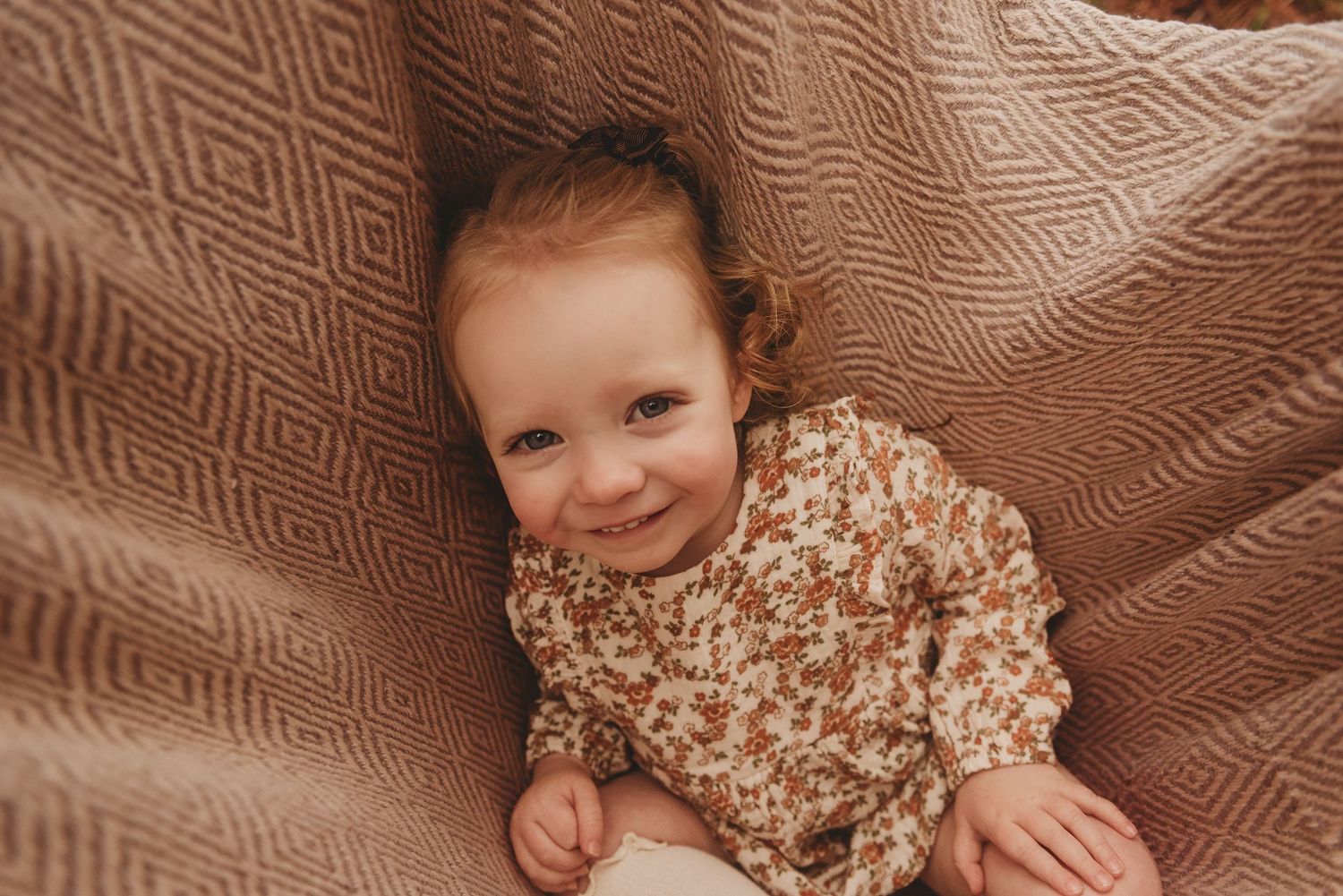 Child smiles while lying on textured brown blanket during outdoor photo session.
