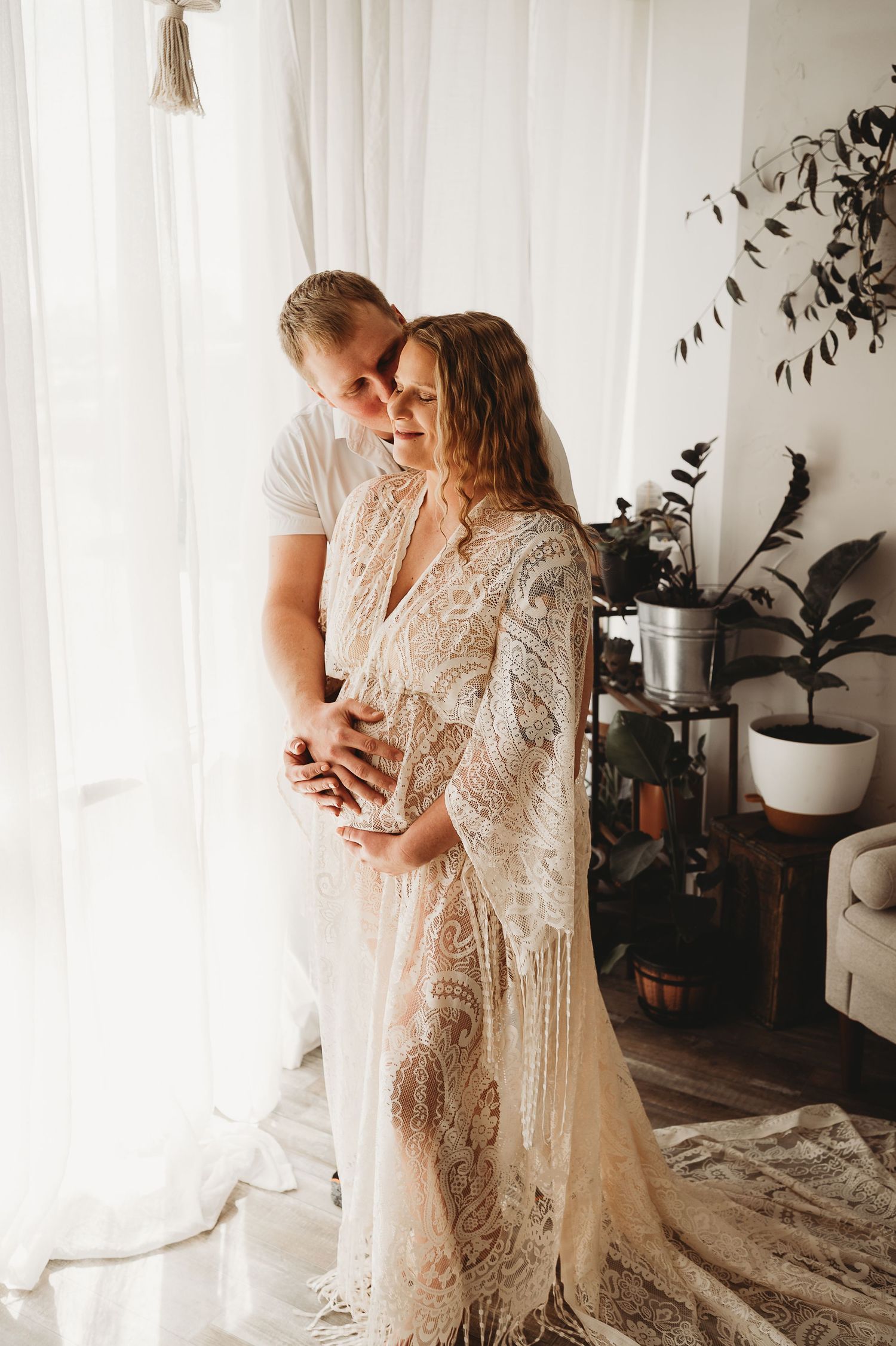 Couple sharing a tender moment by a sunlit window, woman wearing an elegant boho lace maternity gown.