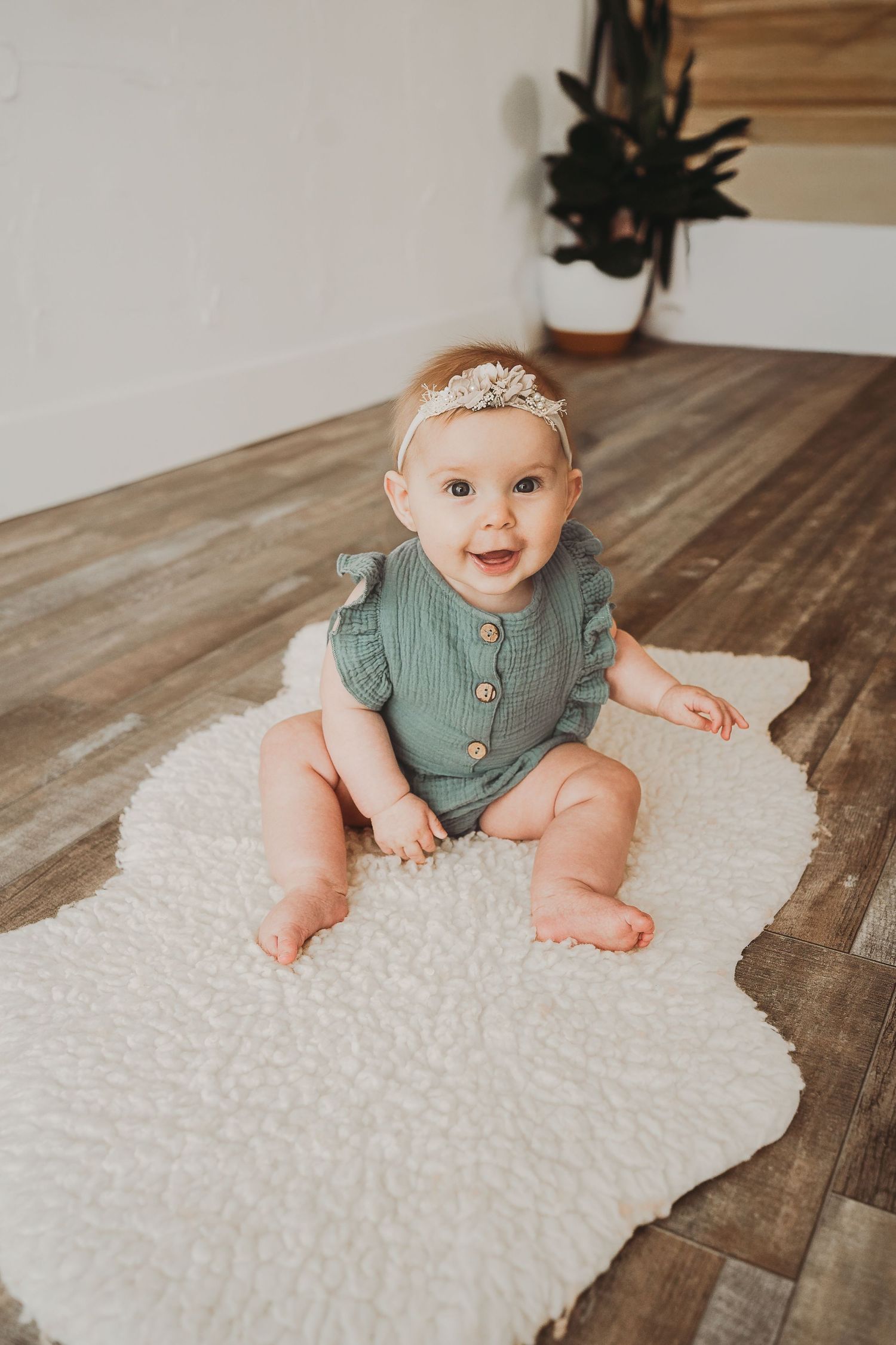Baby in sage green romper sitting on white fluffy rug against wooden floor.