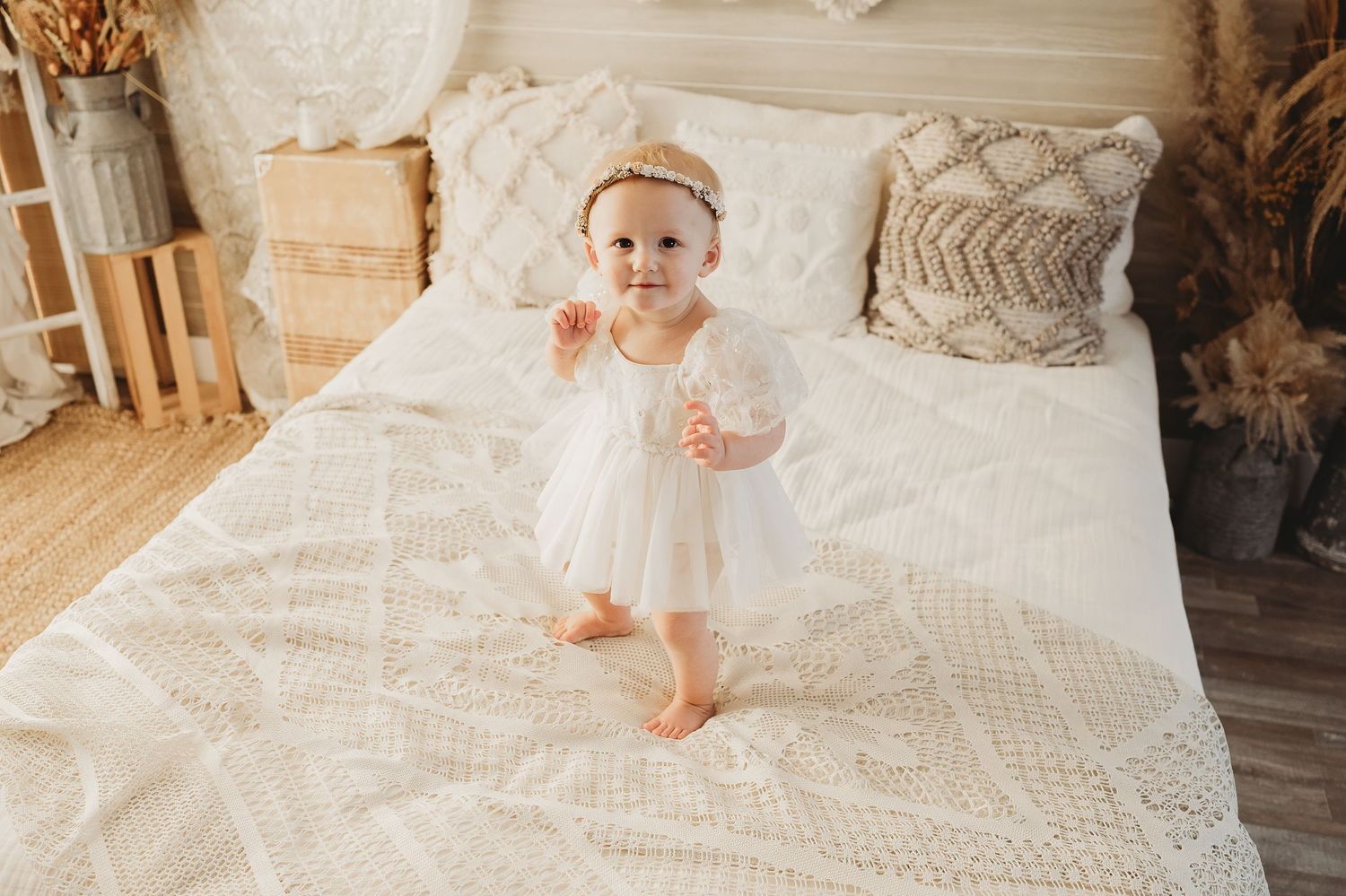 Sweet baby girl in white tutu dress and crown headband stands on knit blanket on bed.