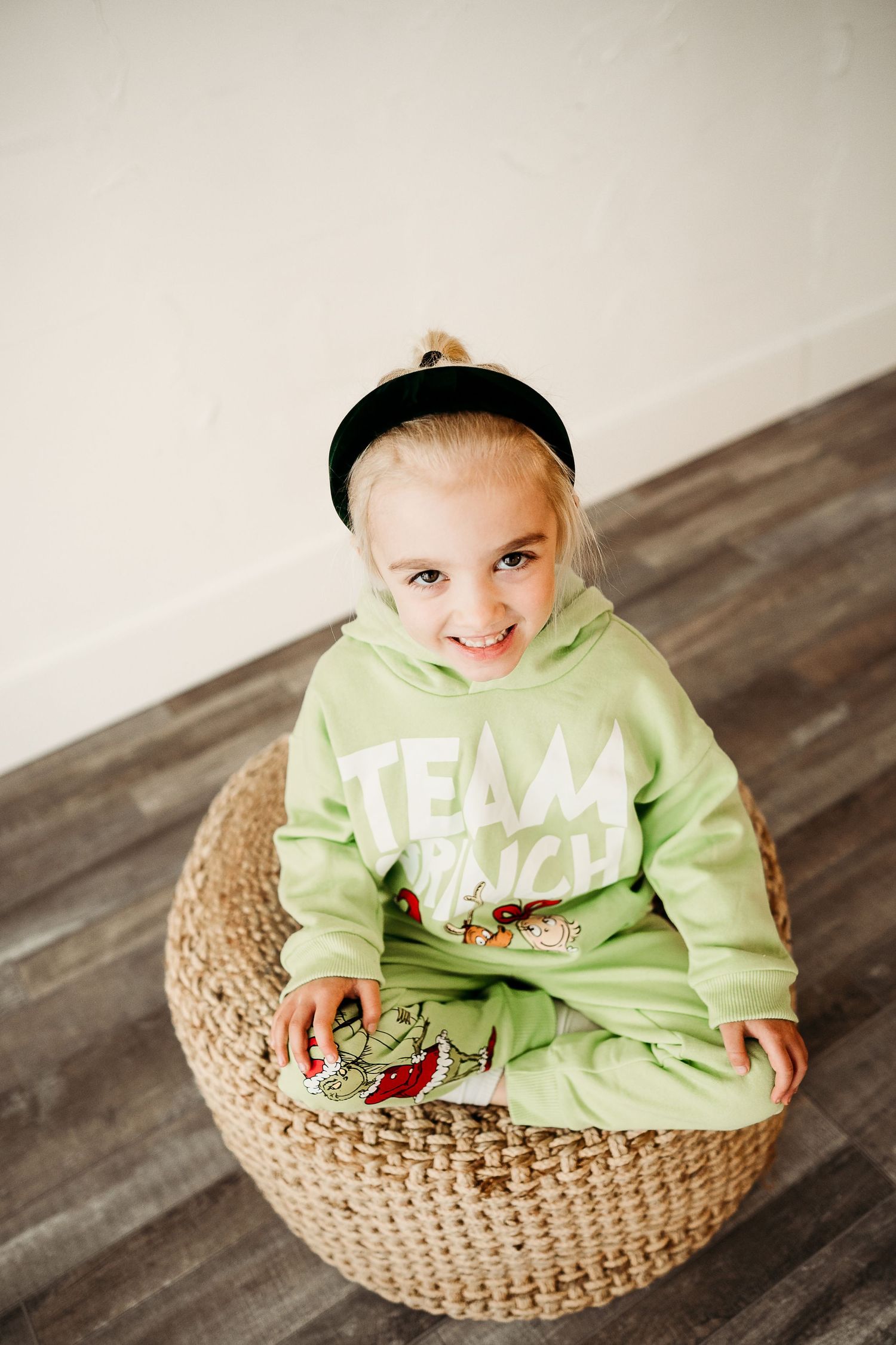 Adorable blonde toddler in green Team Grinch hoodie sits smiling on wicker pouf indoors.