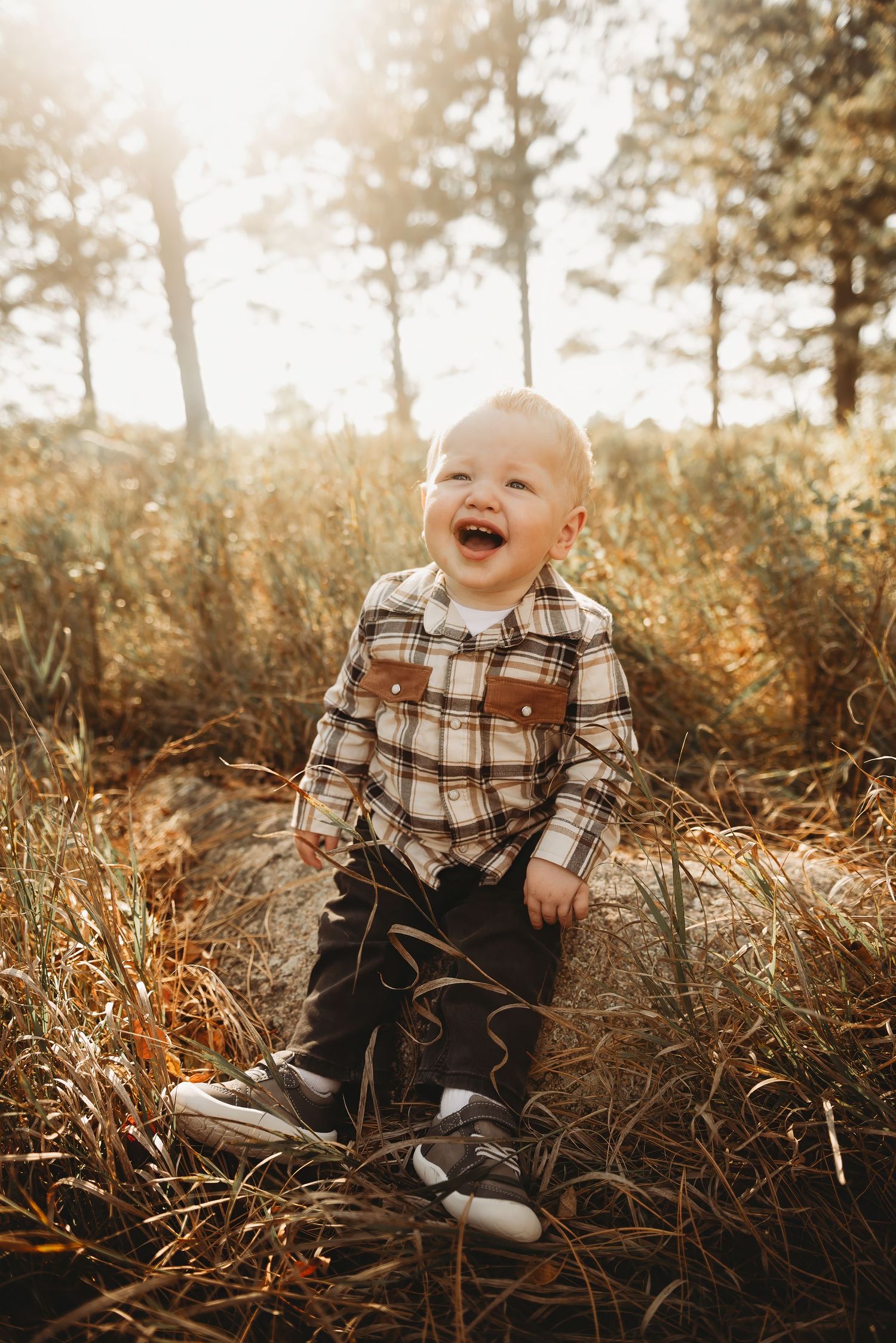 Happy baby in plaid shirt sitting among tall grass during golden hour.
