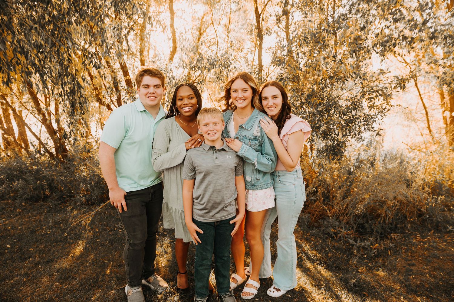 Five smiling young people pose together outdoors among golden-lit trees in a warm family portrait.