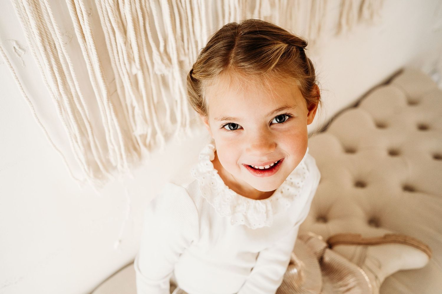 Child in white dress sitting on beige blanket with macrame wall hanging in background.