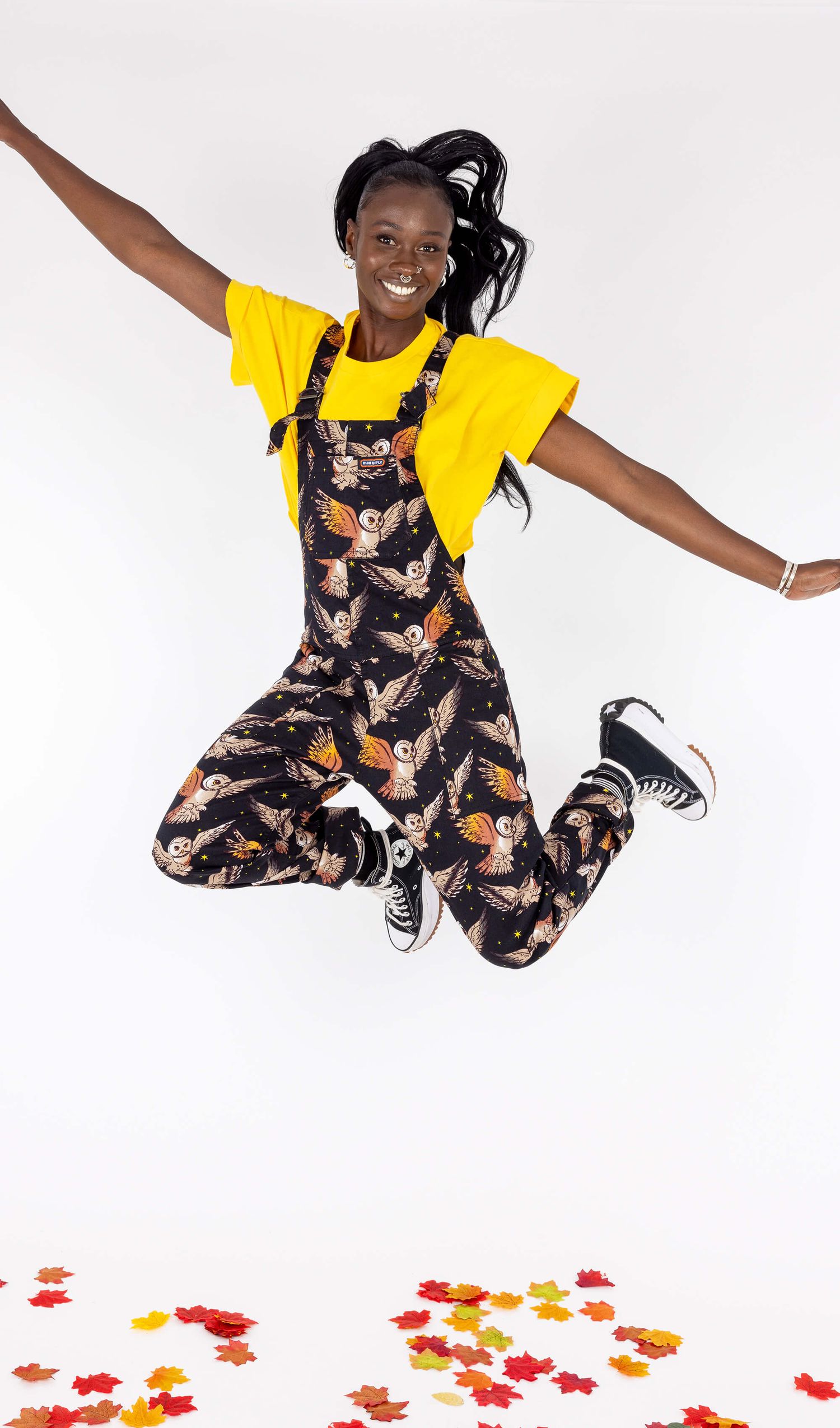 Alternative black model wearing small slow fashion brand Run & Fly owl dungarees with a yellow t-shirt jumping and smili